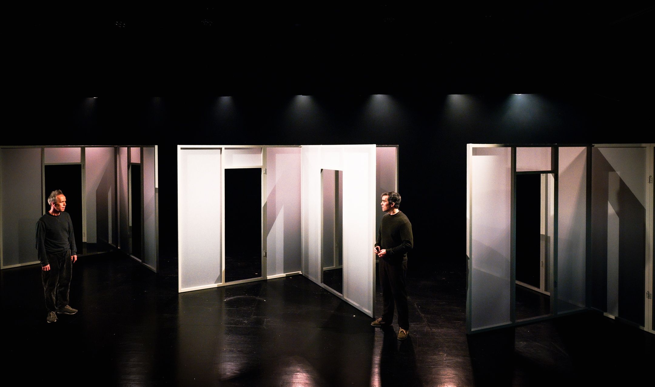 To menn står på hver sin side av en scenografi med hvite panelvegger og åpne døråpninger i et mørkt scenerom.