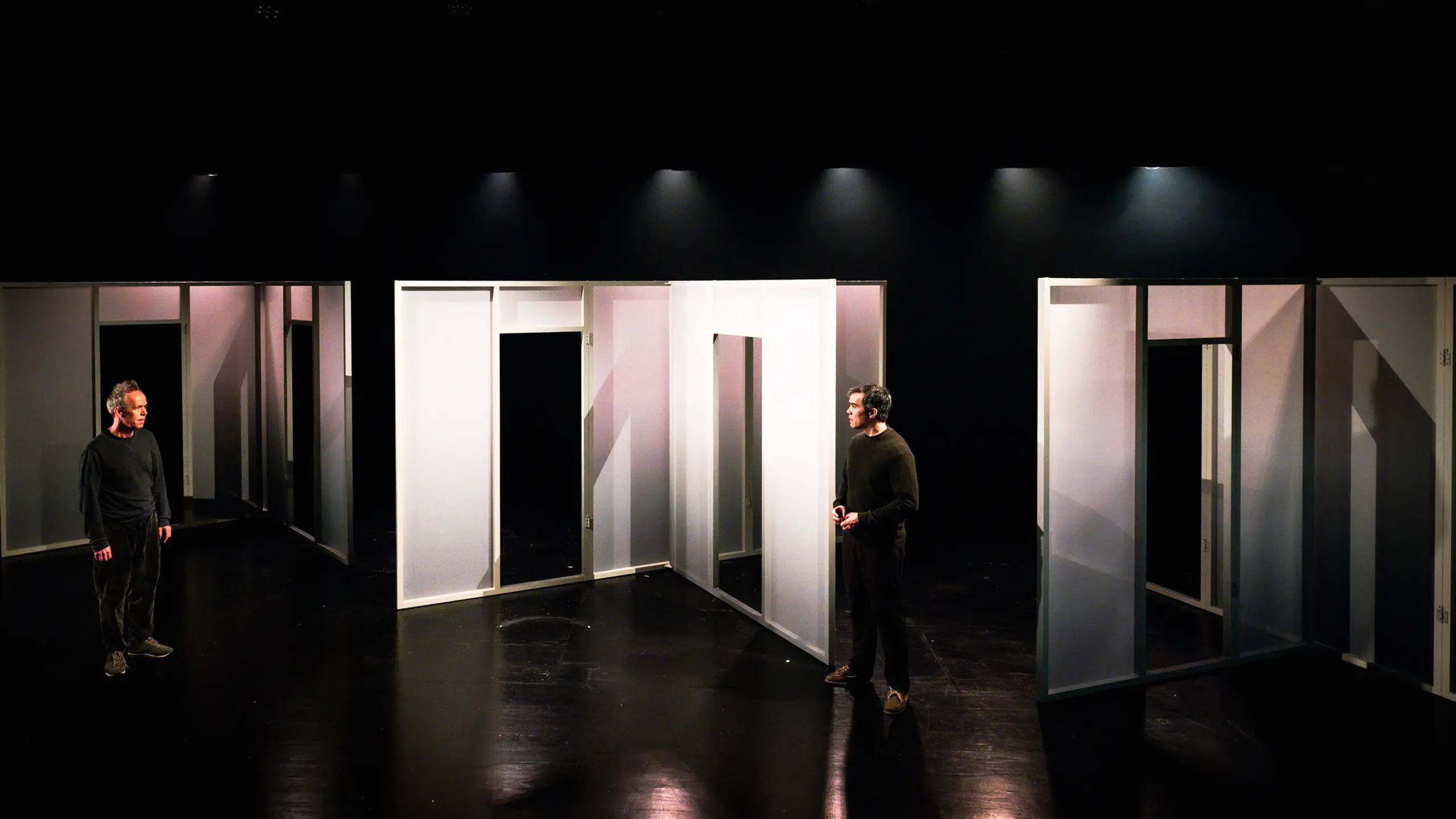 To menn står på hver sin side av en scenografi med hvite panelvegger og åpne døråpninger i et mørkt scenerom.