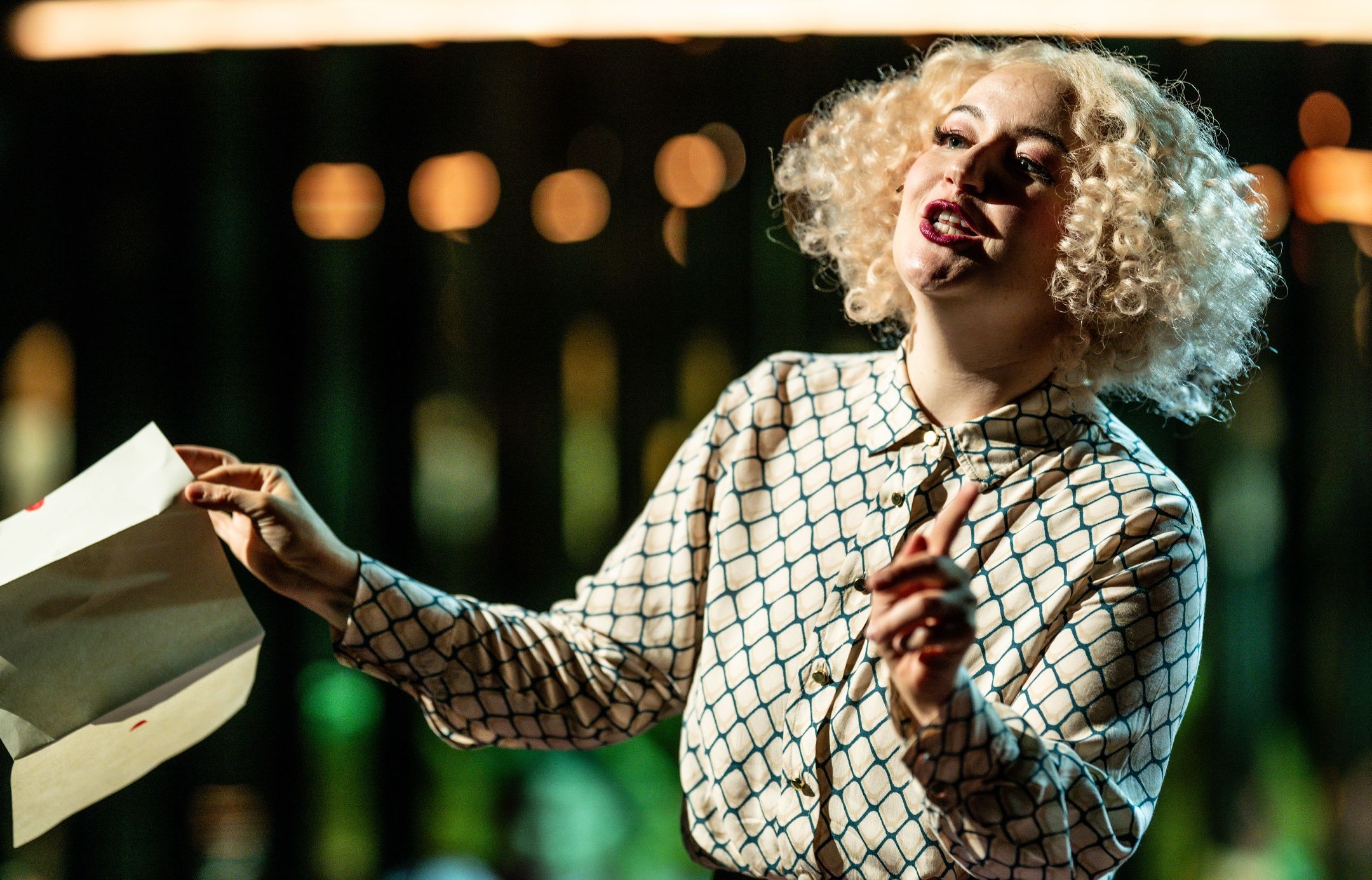 En kvinne med lyst krøllete hår og mønstret skjorte gestikulerer med en konvolutt i hånden.