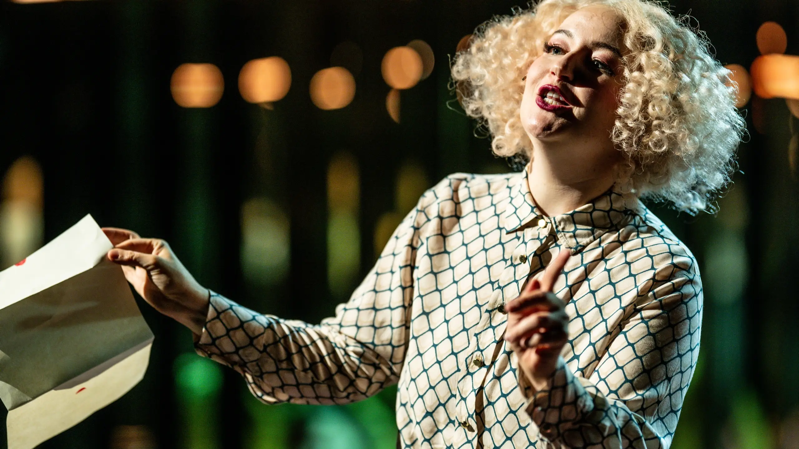 En kvinne med lyst krøllete hår og mønstret skjorte gestikulerer med en konvolutt i hånden.