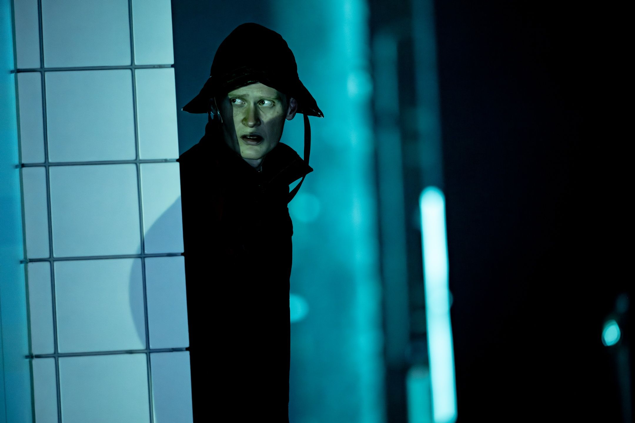 Skuespiller med bøttehatt og mørk jakke kikker rundt et hjørne i blålig scenelys.
