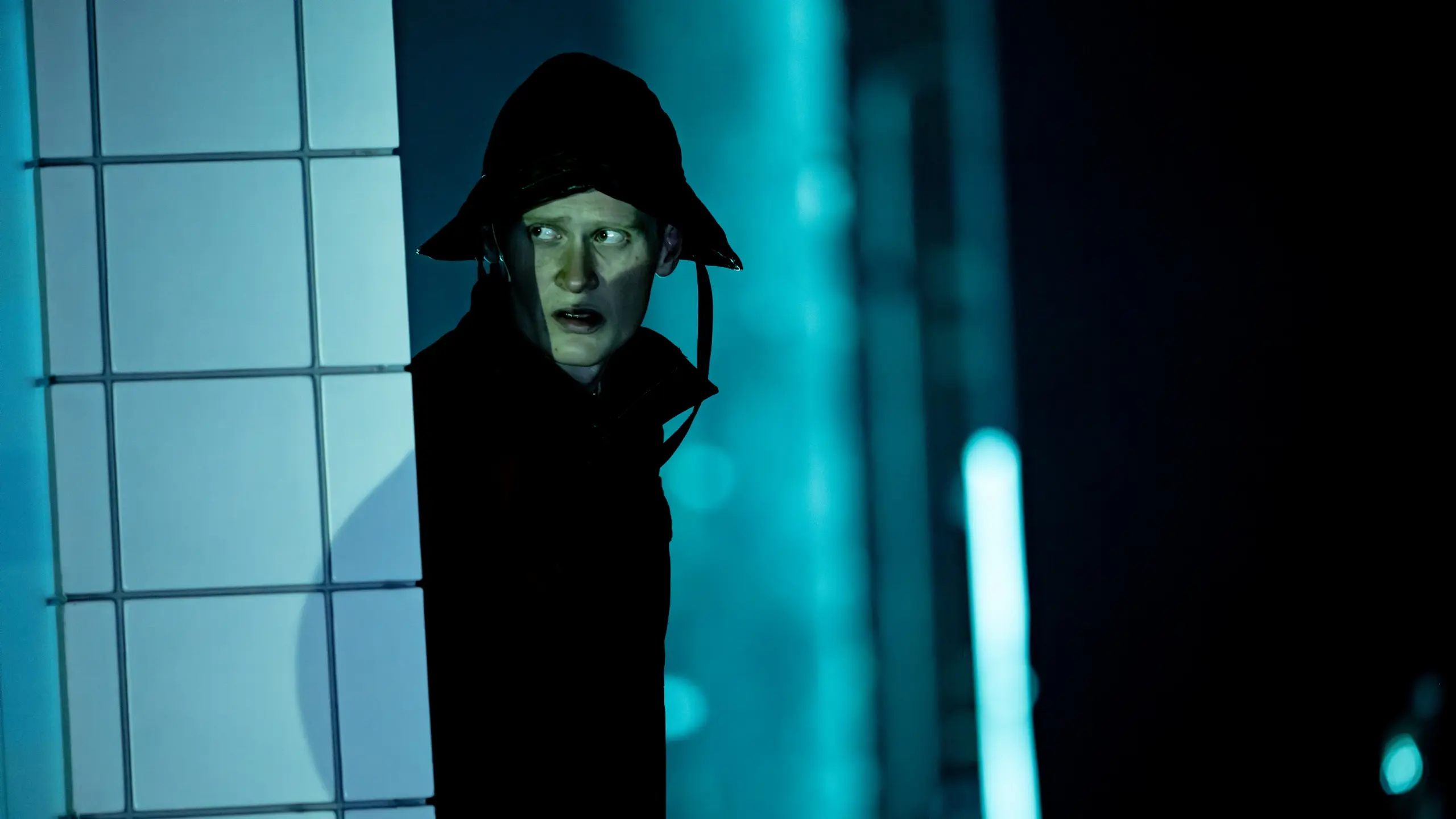 Skuespiller med bøttehatt og mørk jakke kikker rundt et hjørne i blålig scenelys.