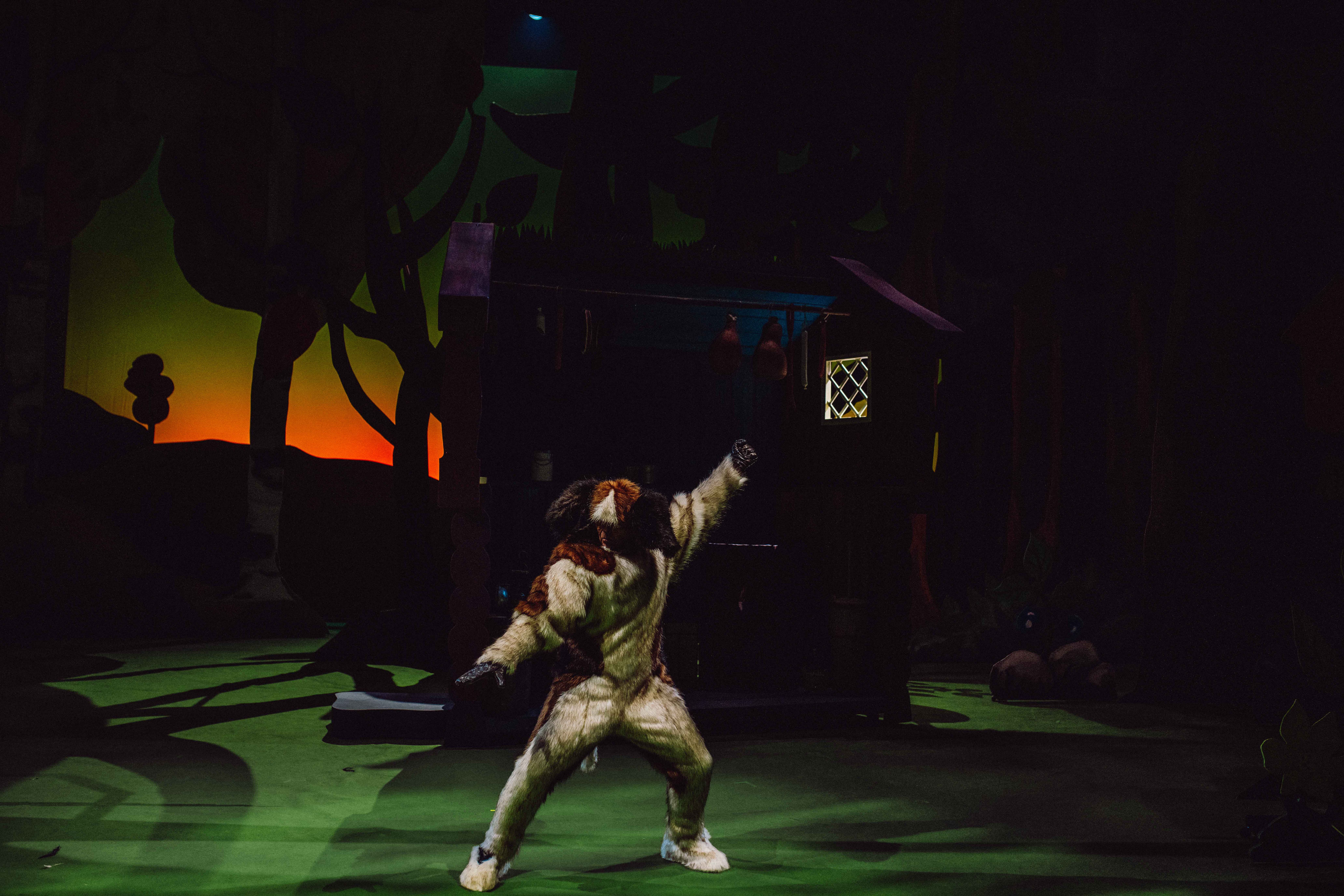 En person i hundekostyme står i en dynamisk positur foran en hytte i kveldsmørket.
