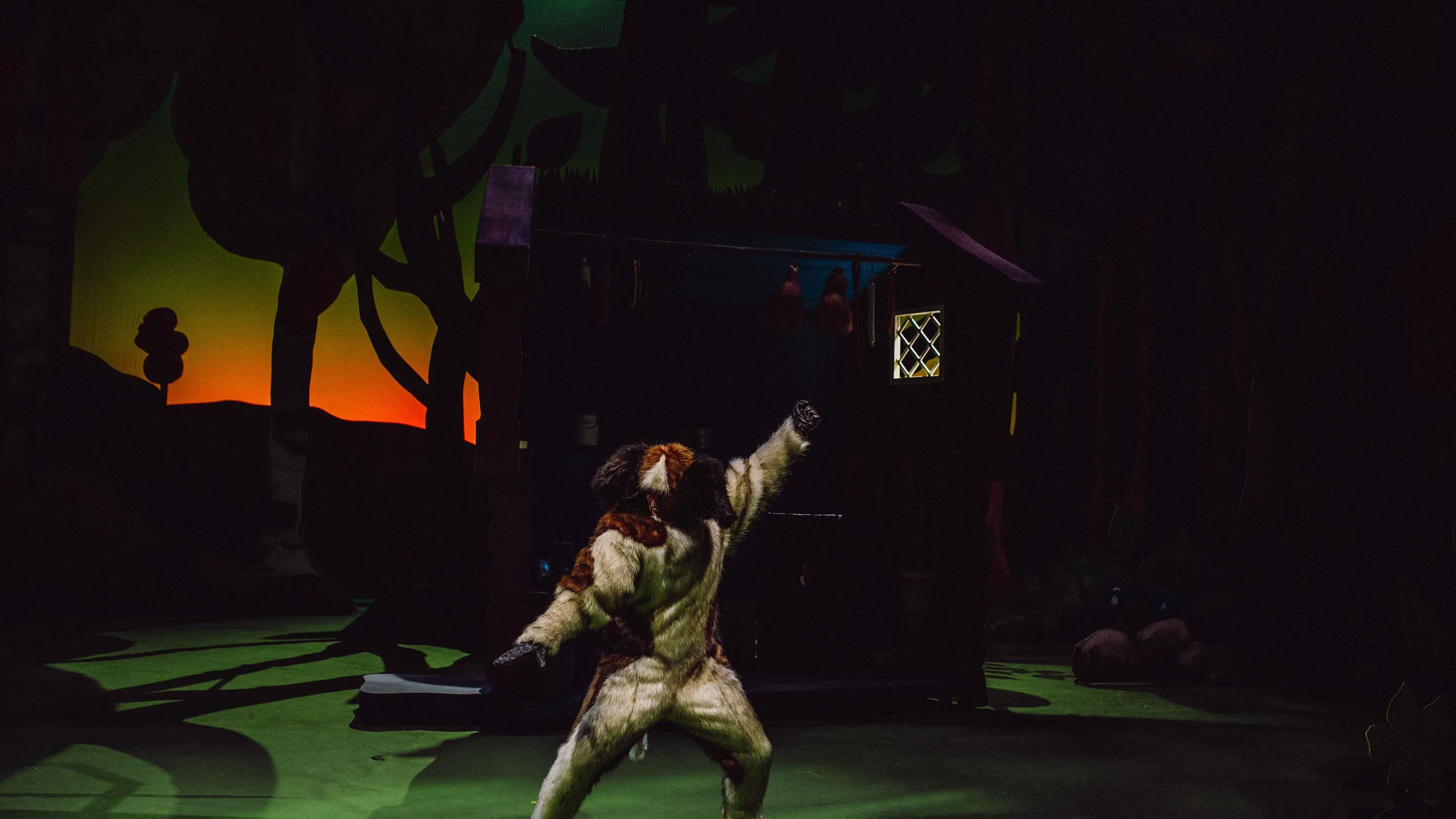 En person i hundekostyme står i en dynamisk positur foran en hytte i kveldsmørket.