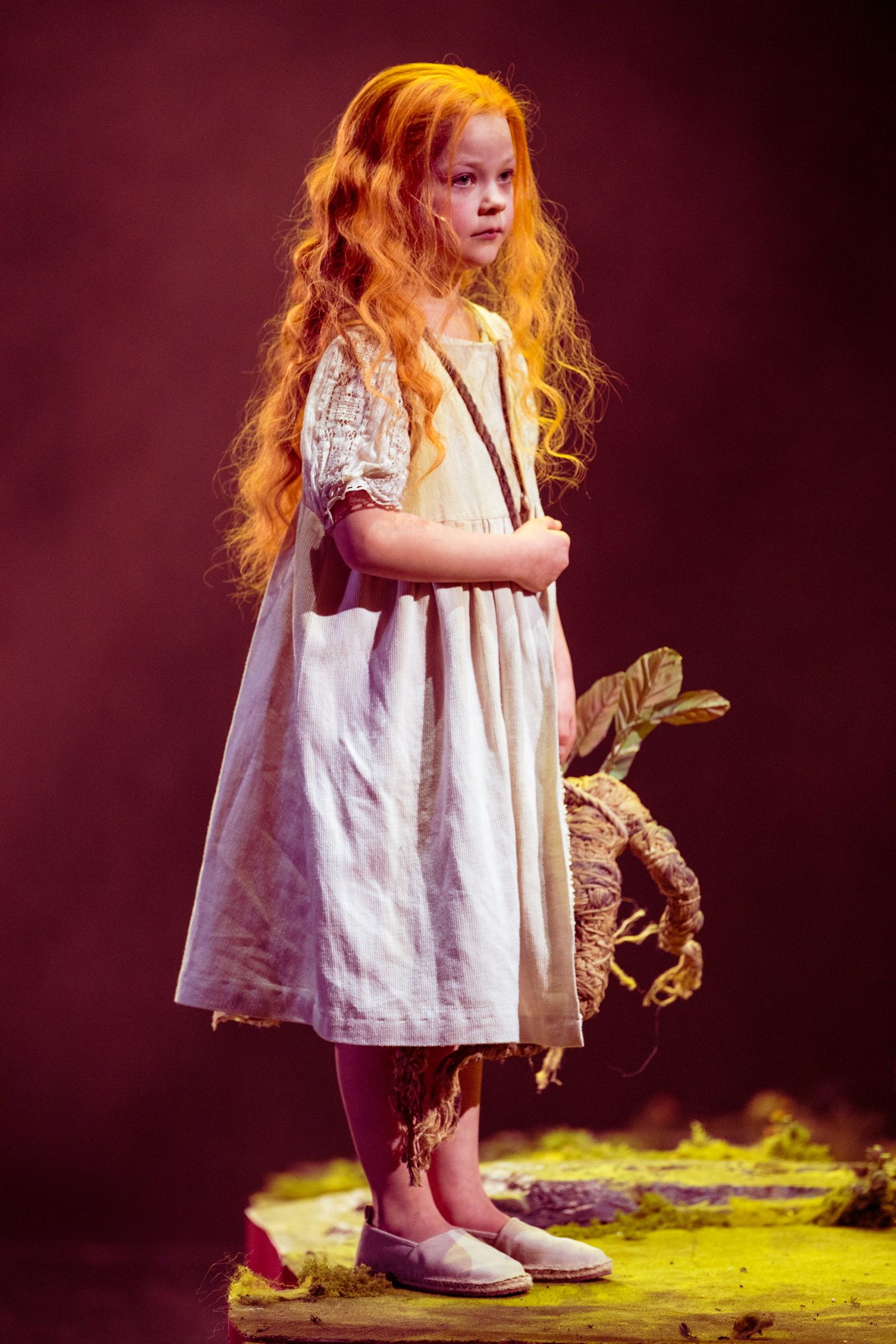 En liten jente med langt, bølgete rødt hår og lys kjole står alvorlig på scenen med en dukke av tau og blader i hånden.