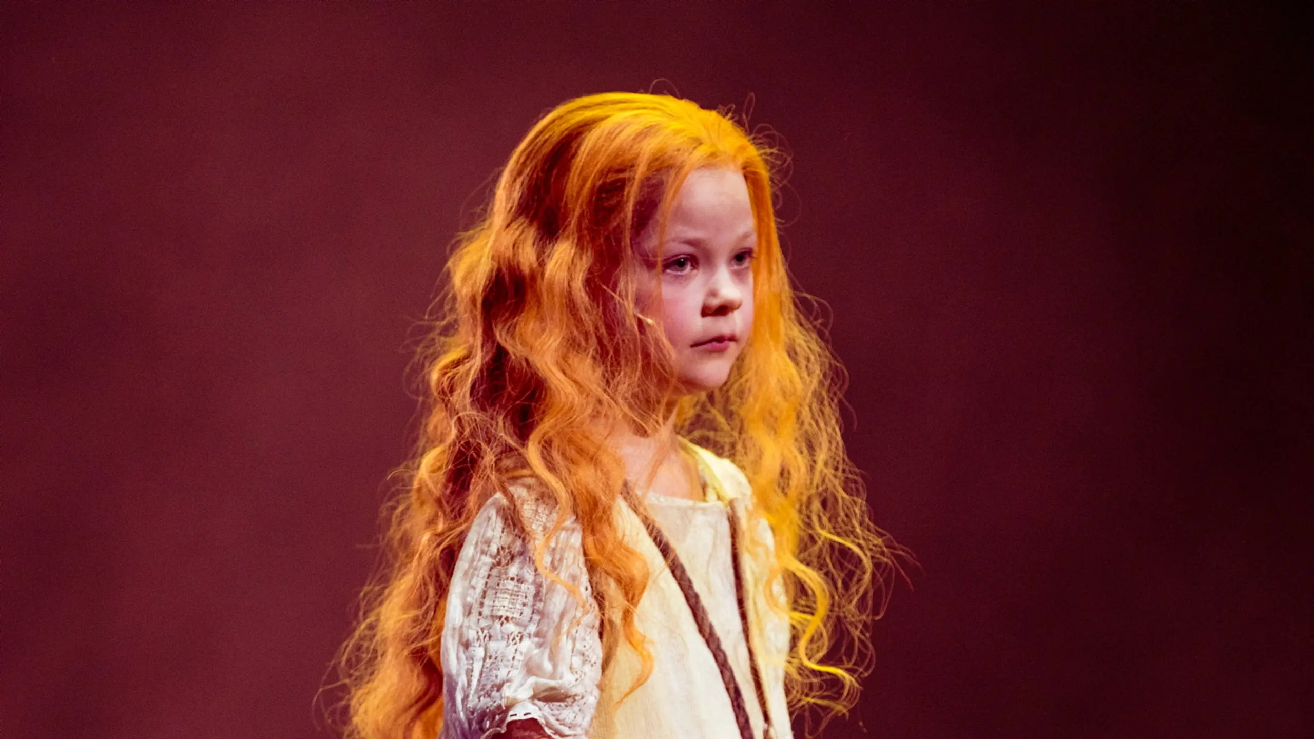 En liten jente med langt, bølgete rødt hår og lys kjole står alvorlig på scenen med en dukke av tau og blader i hånden.