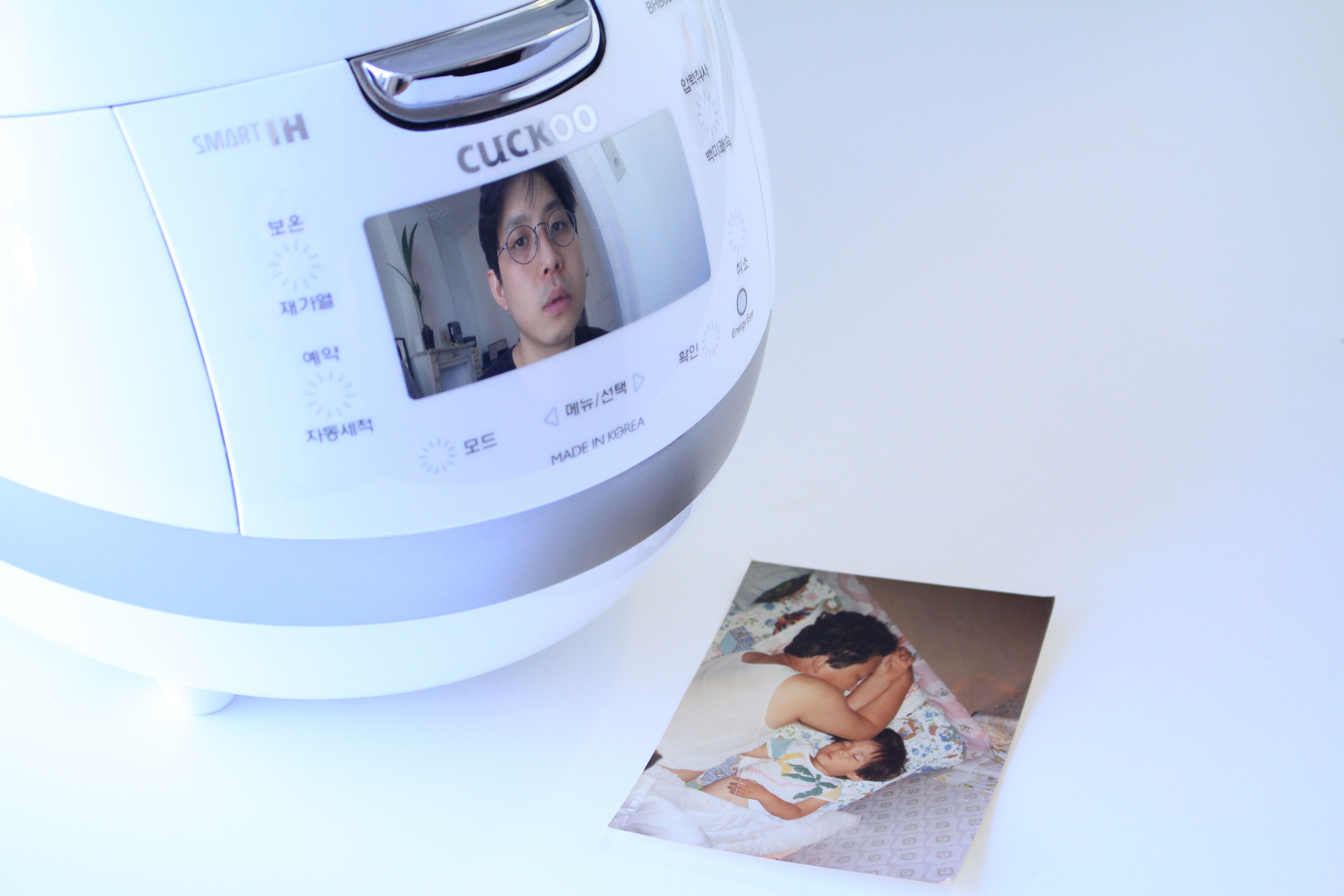 Et bilde av en elektrisk riskoker, hvor det vises bilde av en ung sørkoreansk mann på skjermen. Ved siden av riskokeren ligger det et fotografi av en voksen mann som sover ved siden av et barn.