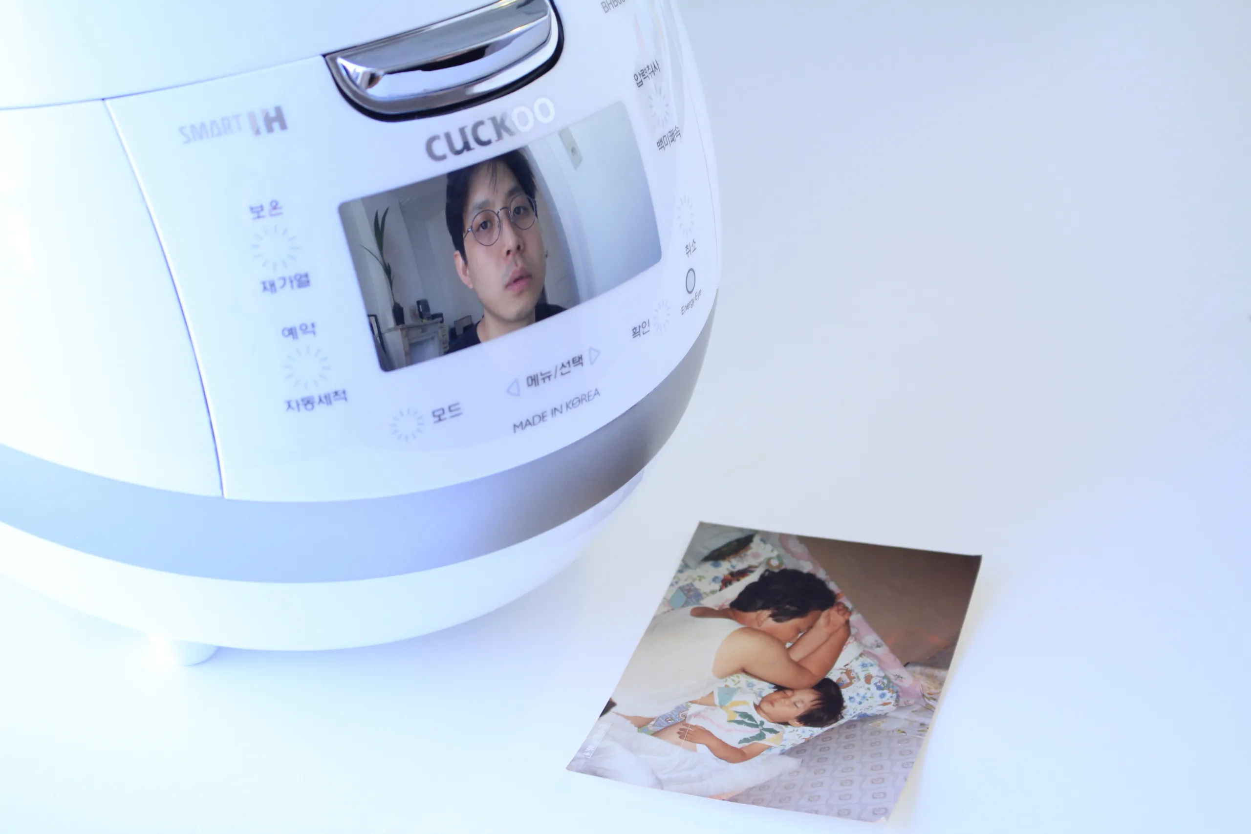 Et bilde av en elektrisk riskoker, hvor det vises bilde av en ung sørkoreansk mann på skjermen. Ved siden av riskokeren ligger det et fotografi av en voksen mann som sover ved siden av et barn.