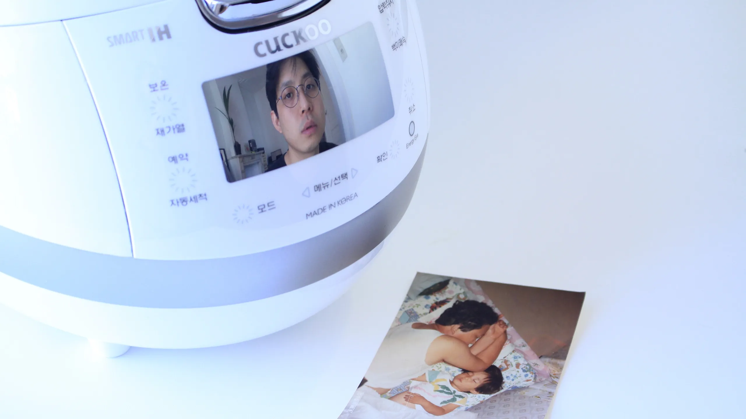 Et bilde av en elektrisk riskoker, hvor det vises bilde av en ung sørkoreansk mann på skjermen. Ved siden av riskokeren ligger det et fotografi av en voksen mann som sover ved siden av et barn.