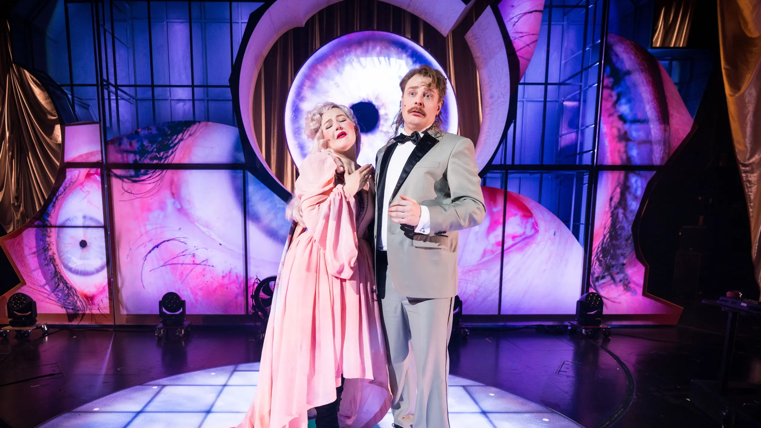 To skuespillere står tett sammen på en teaterscene. Personen til venstre har på seg en stor rosa kjole med lange ermer og hvit parykk, og holder hånden dramatisk til brystet. Personen til høyre har på seg en lys grå dress. Bak dem vises en stor projeksjon av et blått øye