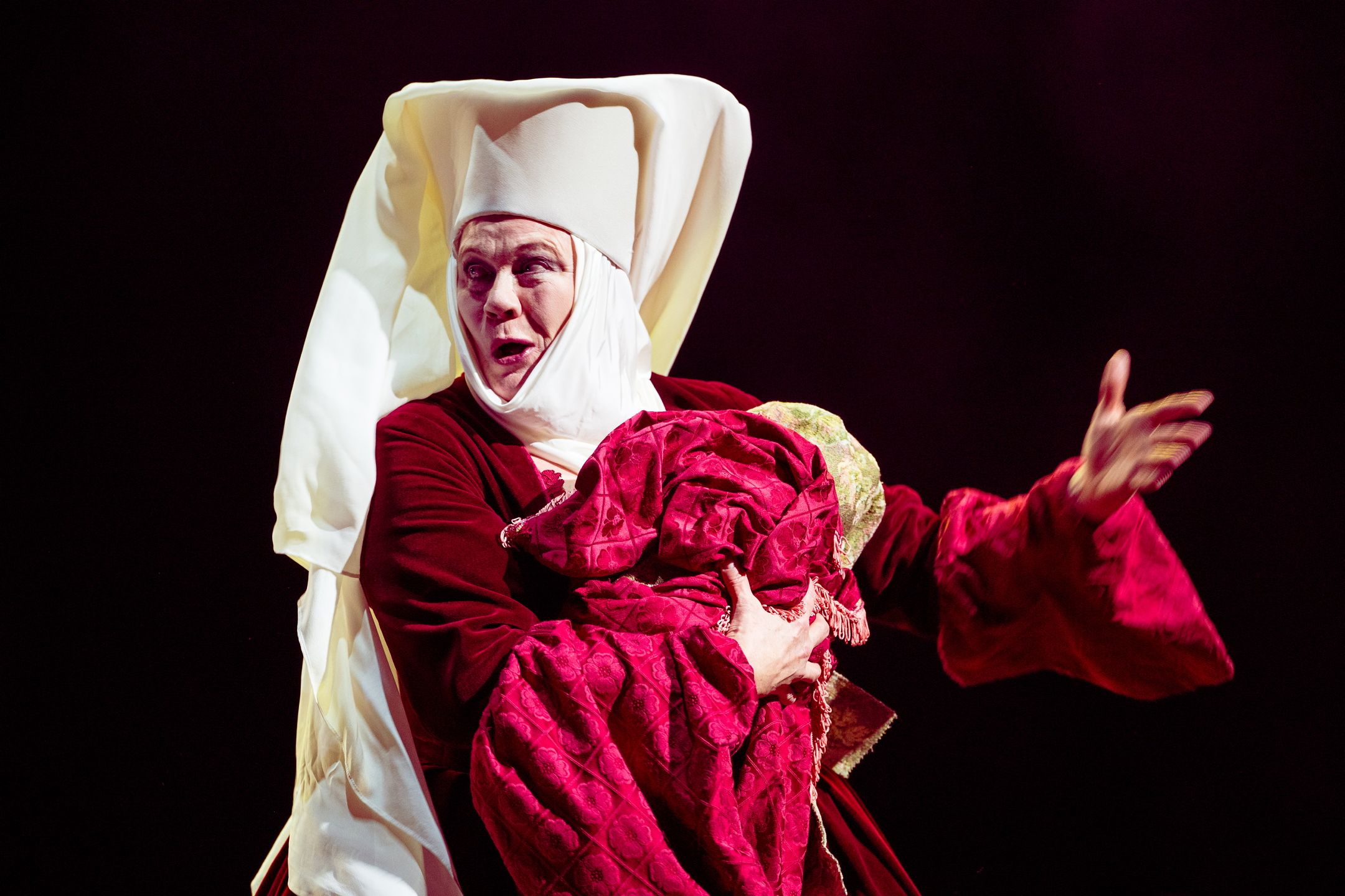 kvinne i rød og hvit nonnedrakt holder en bylt med rødt teppe og gestikulerer dramatisk med fri arm.