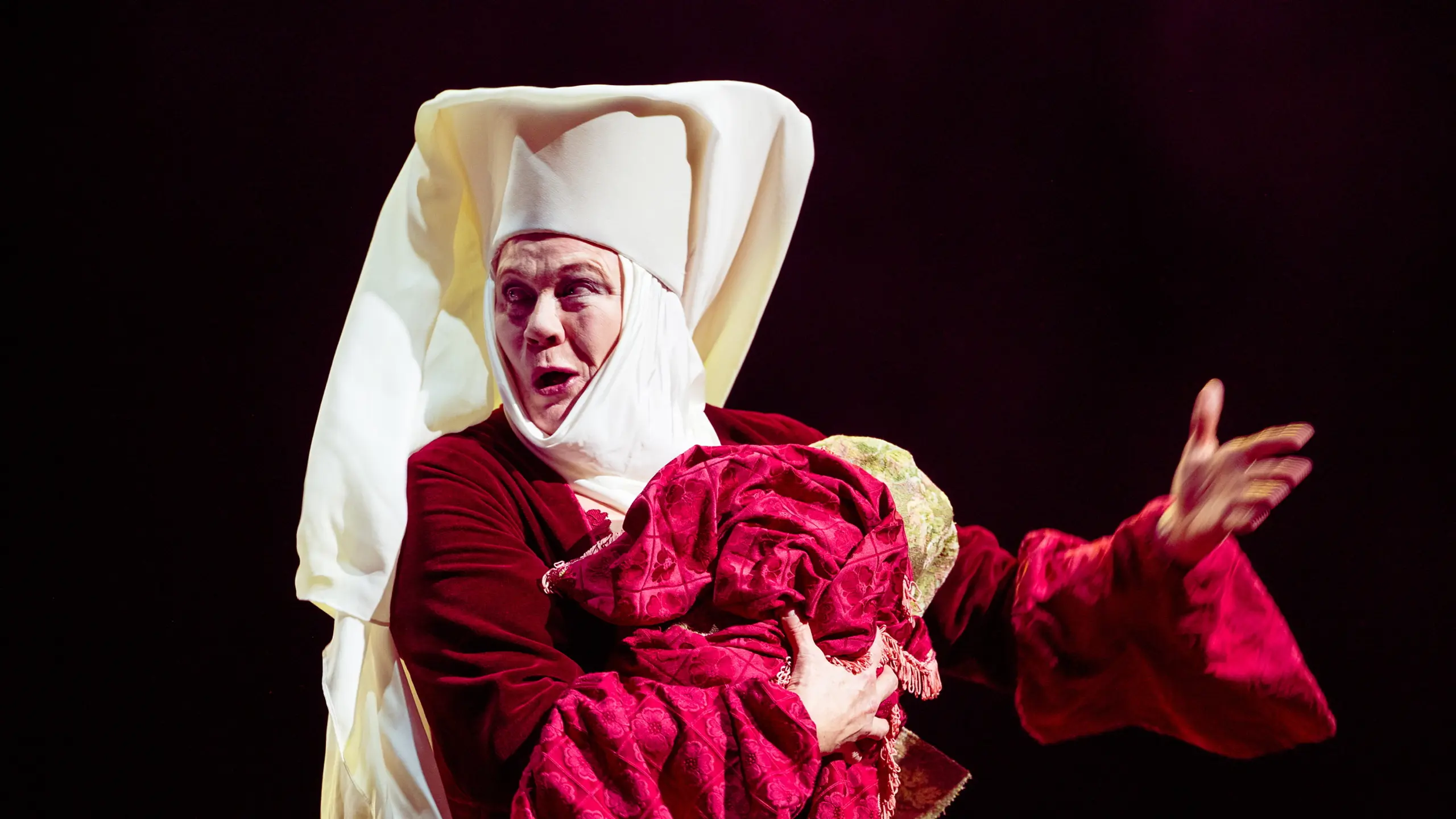 kvinne i rød og hvit nonnedrakt holder en bylt med rødt teppe og gestikulerer dramatisk med fri arm.