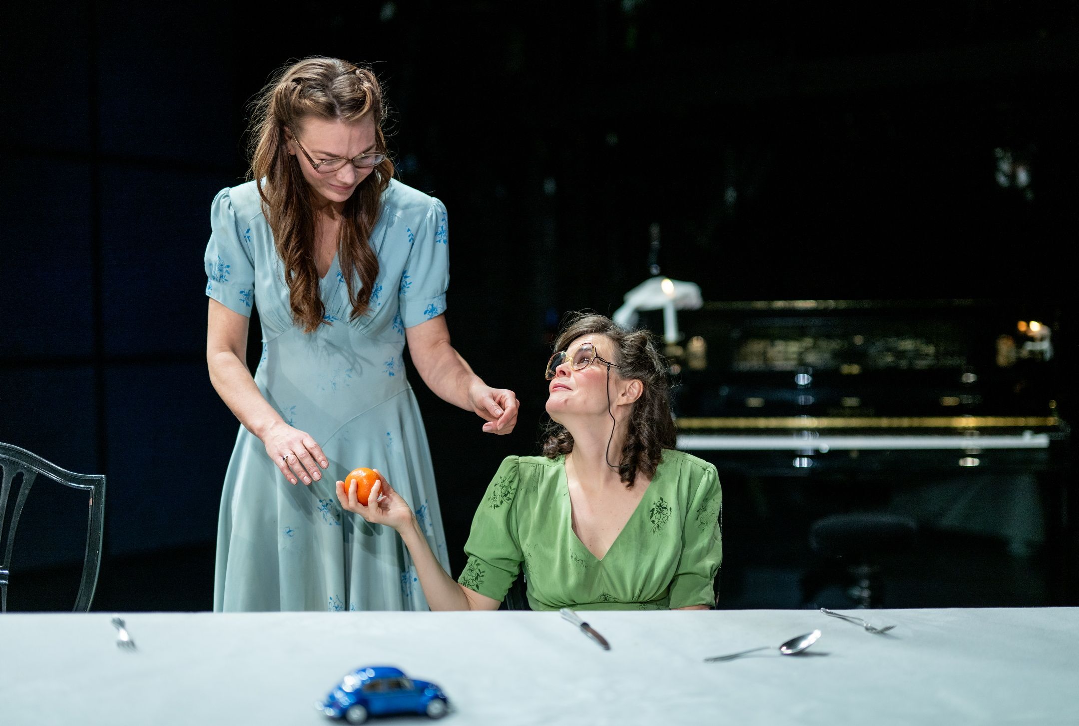 To kvinner i grønne og blå kjoler ved et bord; den stående rekker en klementin til den sittende.