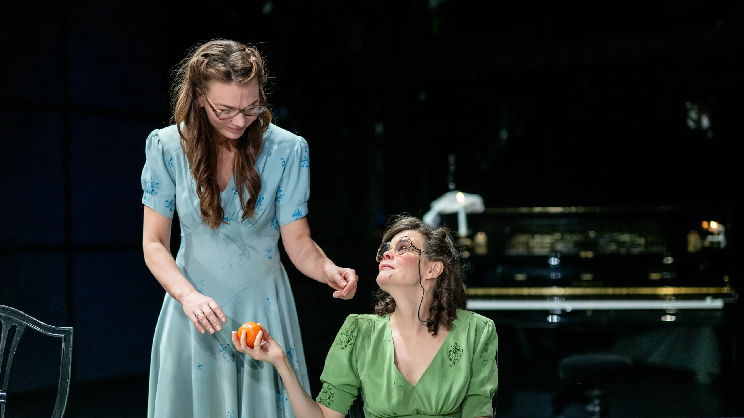 To kvinner i grønne og blå kjoler ved et bord; den stående rekker en klementin til den sittende.