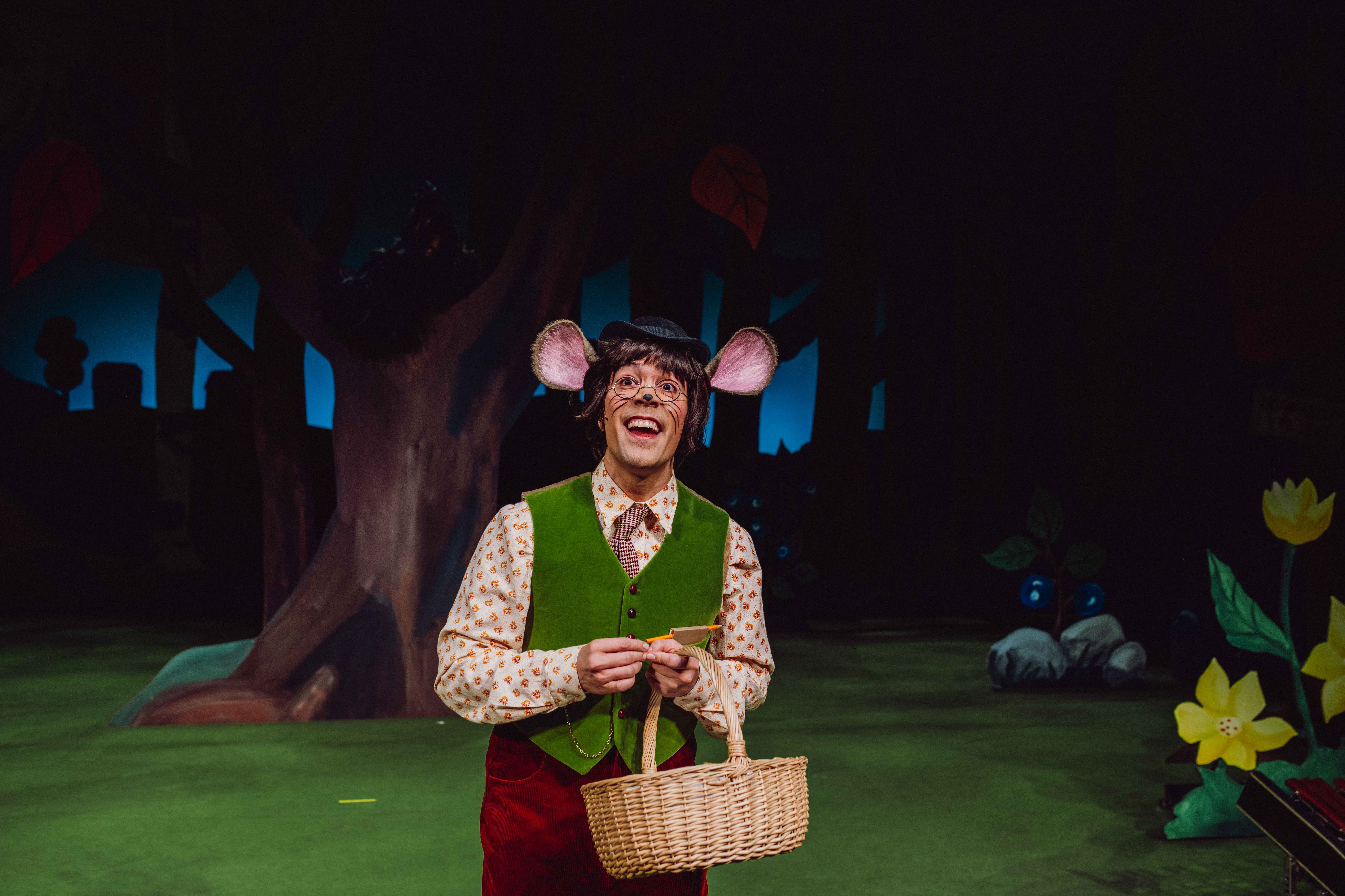 En karakter utkledd som mus med vest og kurv smiler på en skogscene.