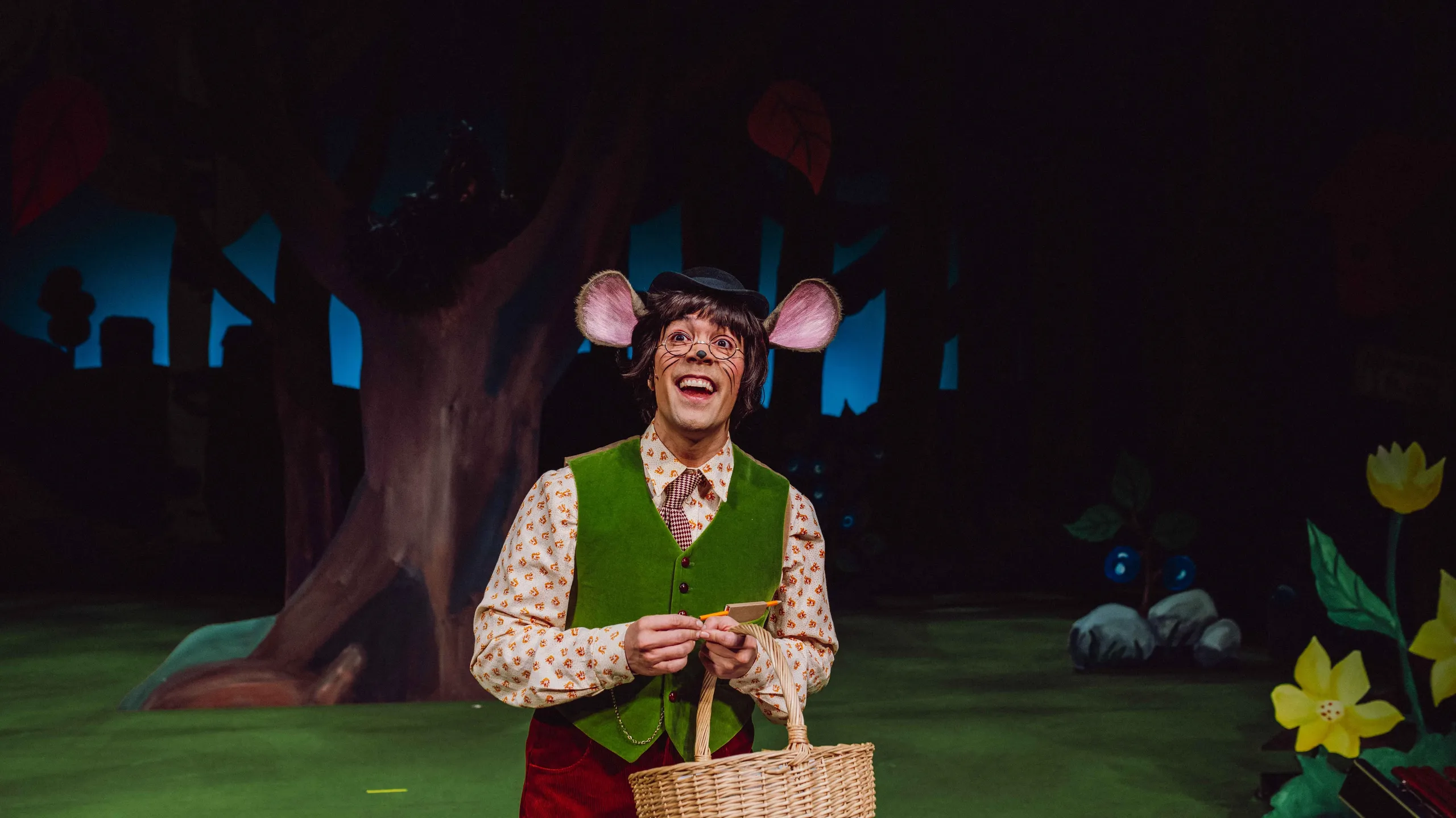 En karakter utkledd som mus med vest og kurv smiler på en skogscene.