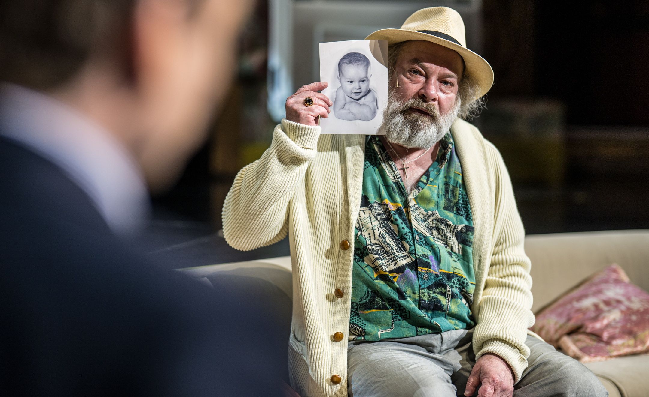 mann med skjegg og hatt holder opp svart-hvitt bilde av baby mens han sitter i sofa