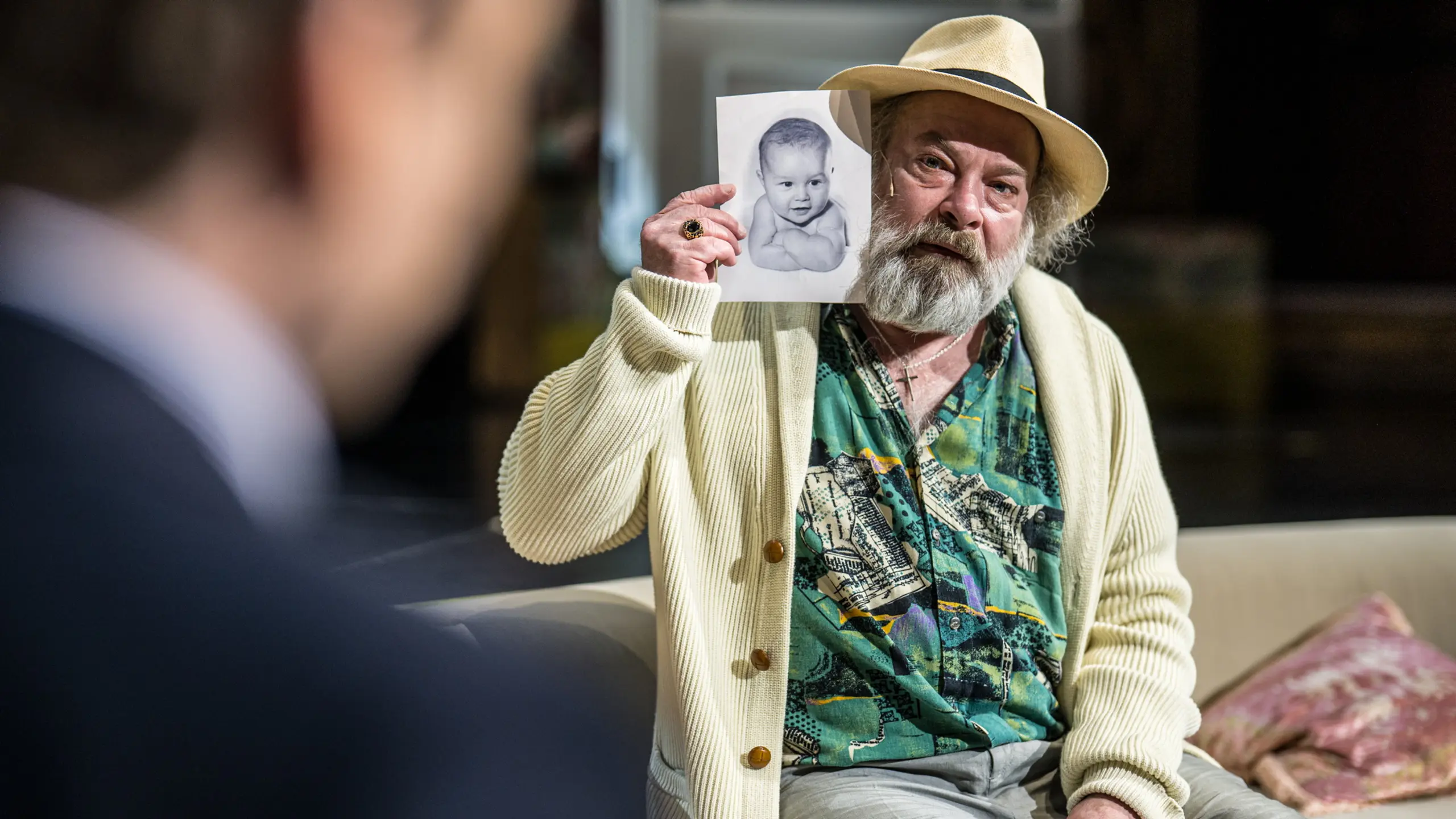 mann med skjegg og hatt holder opp svart-hvitt bilde av baby mens han sitter i sofa