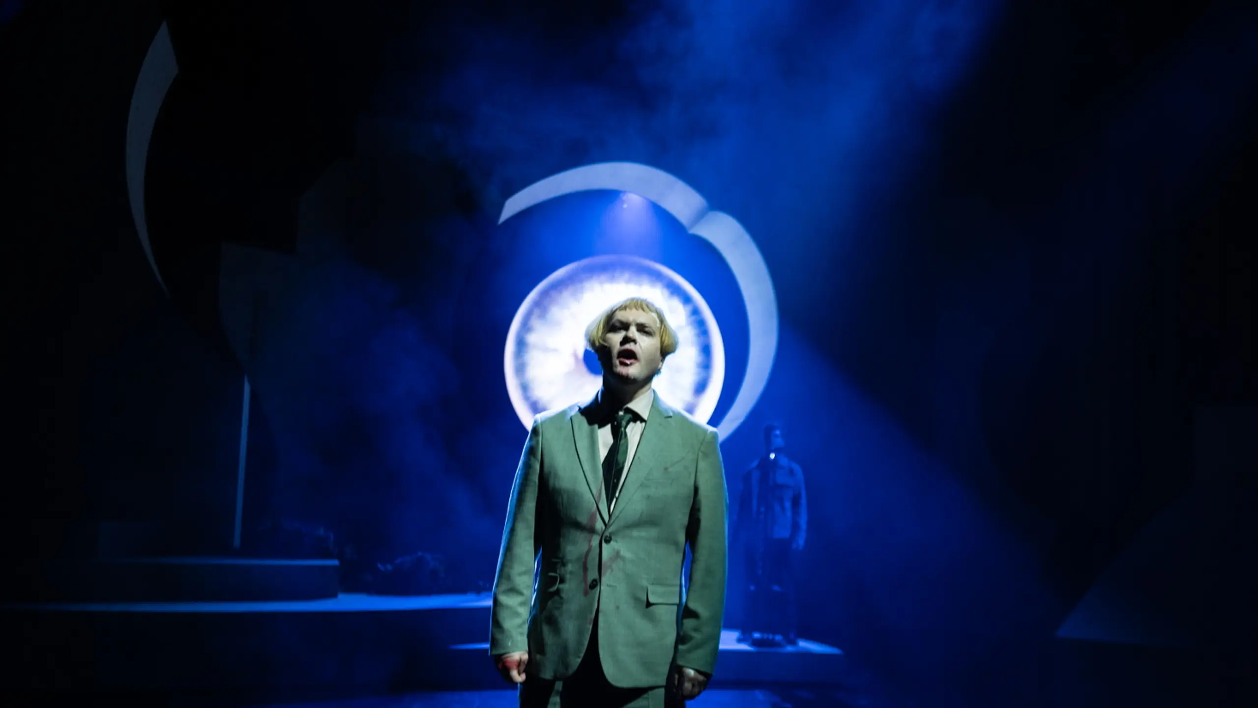 Mann i dress på mørk scene med blått lys. Et stort øye i bakgrunnen