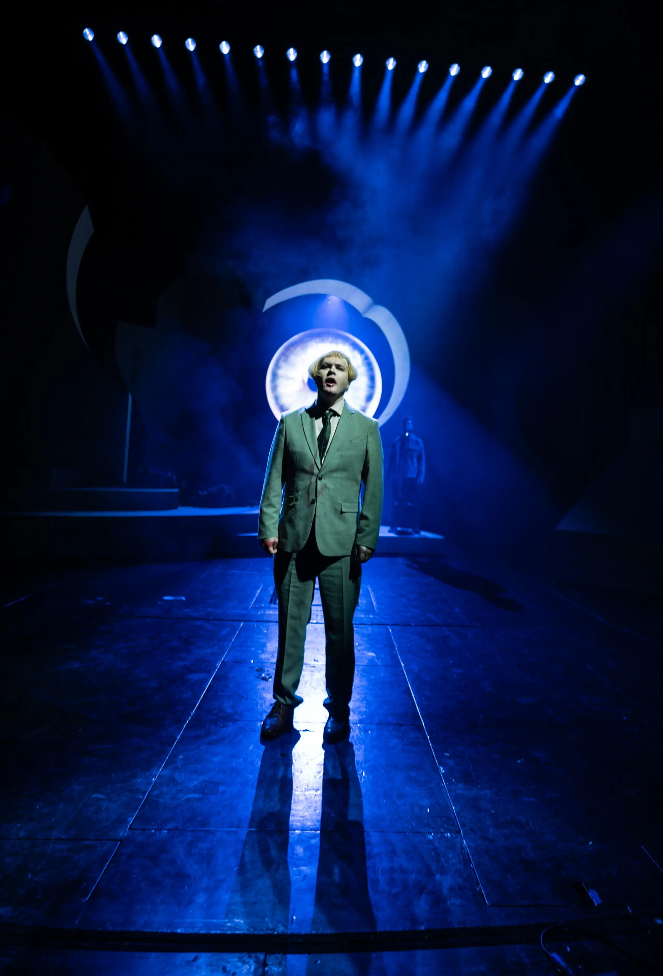 Mann i dress på mørk scene med blått lys. Et stort øye i bakgrunnen