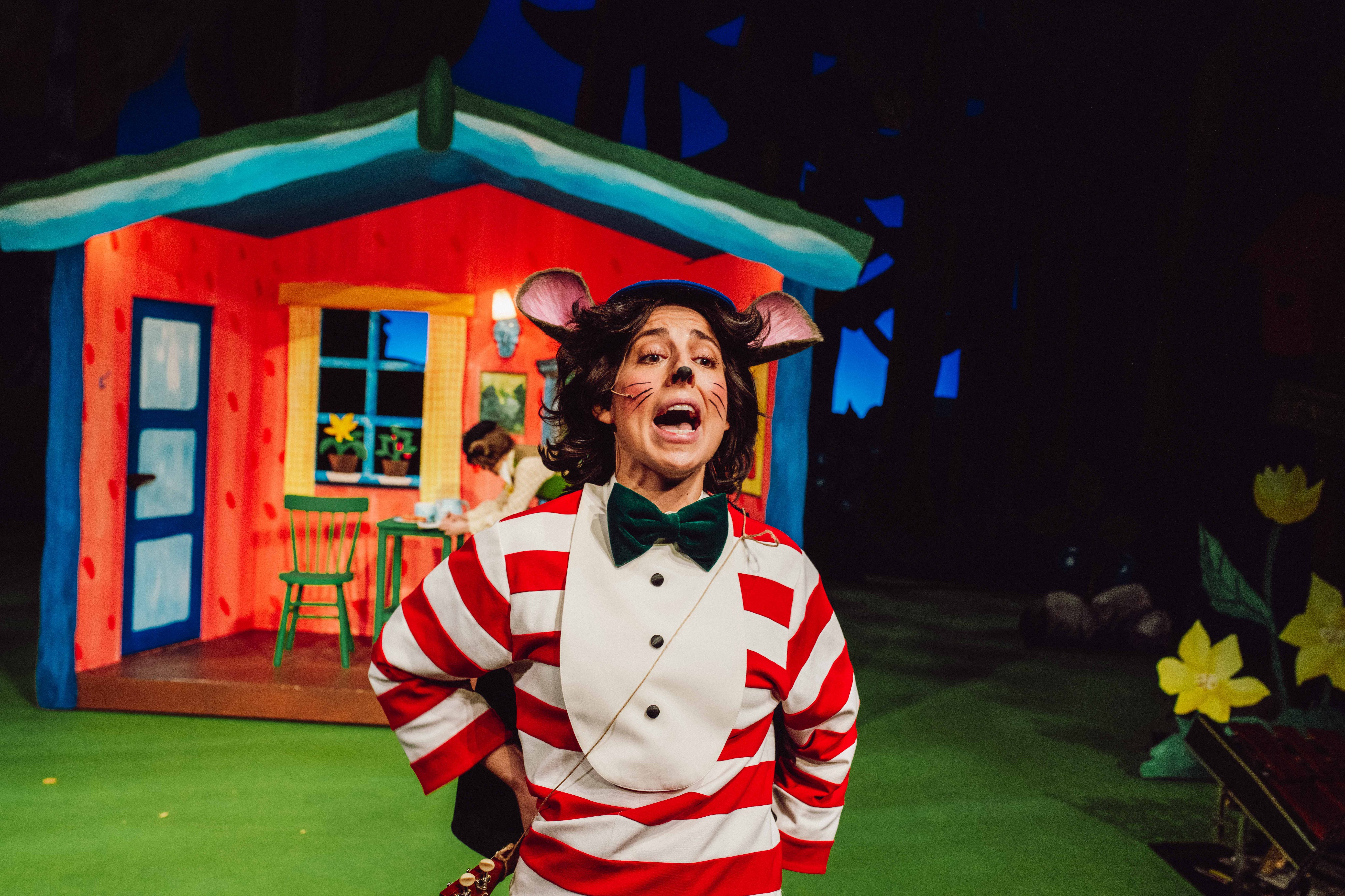 Mus i rød- og hvitstripet genser roper foran fargerik hytte og blomster.