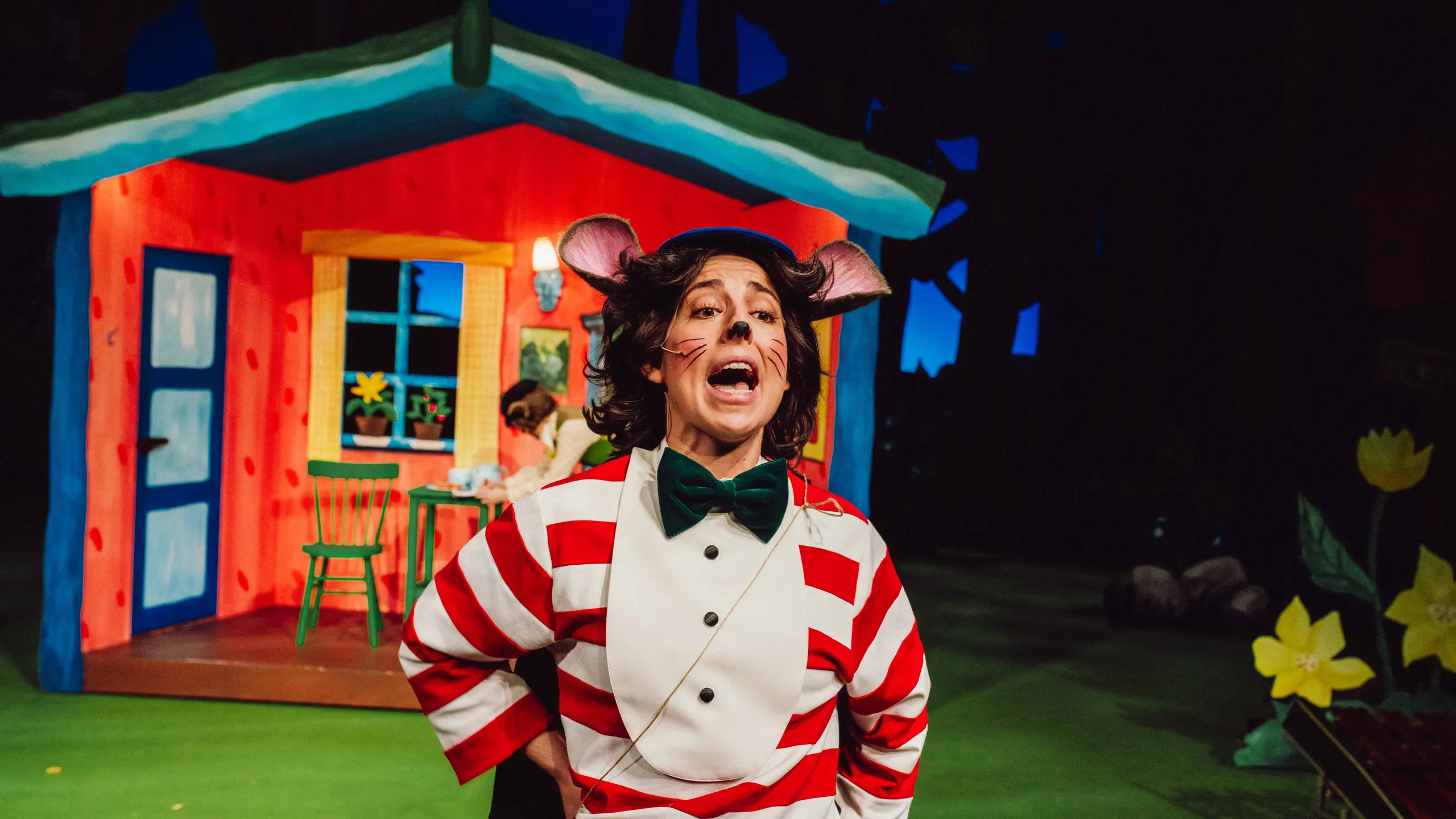 Mus i rød- og hvitstripet genser roper foran fargerik hytte og blomster.