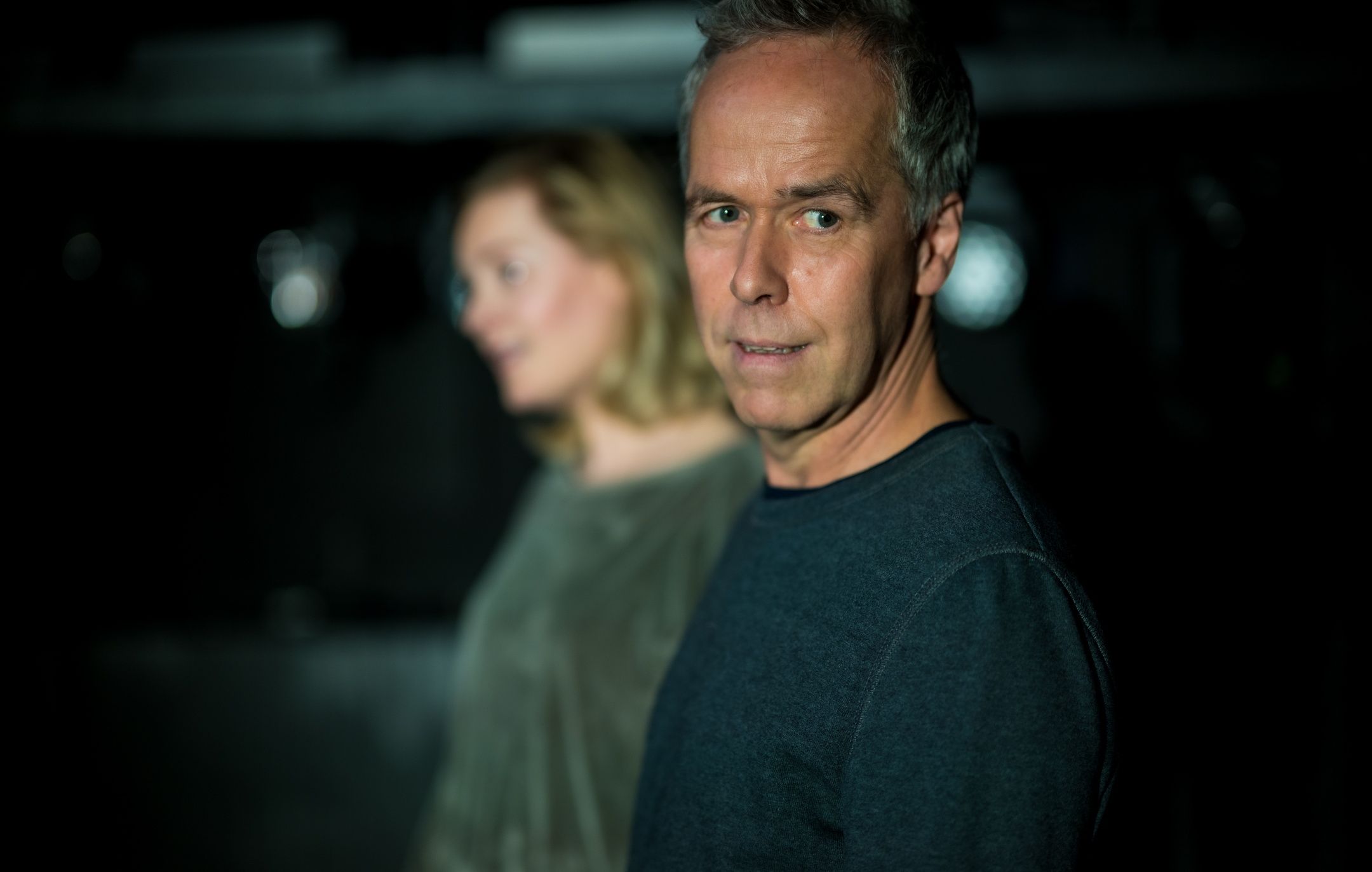 En mann ser rett mot kamera med nøytral mine, mens en kvinne står bak ham i uskarpt fokus, begge i mørke klær.