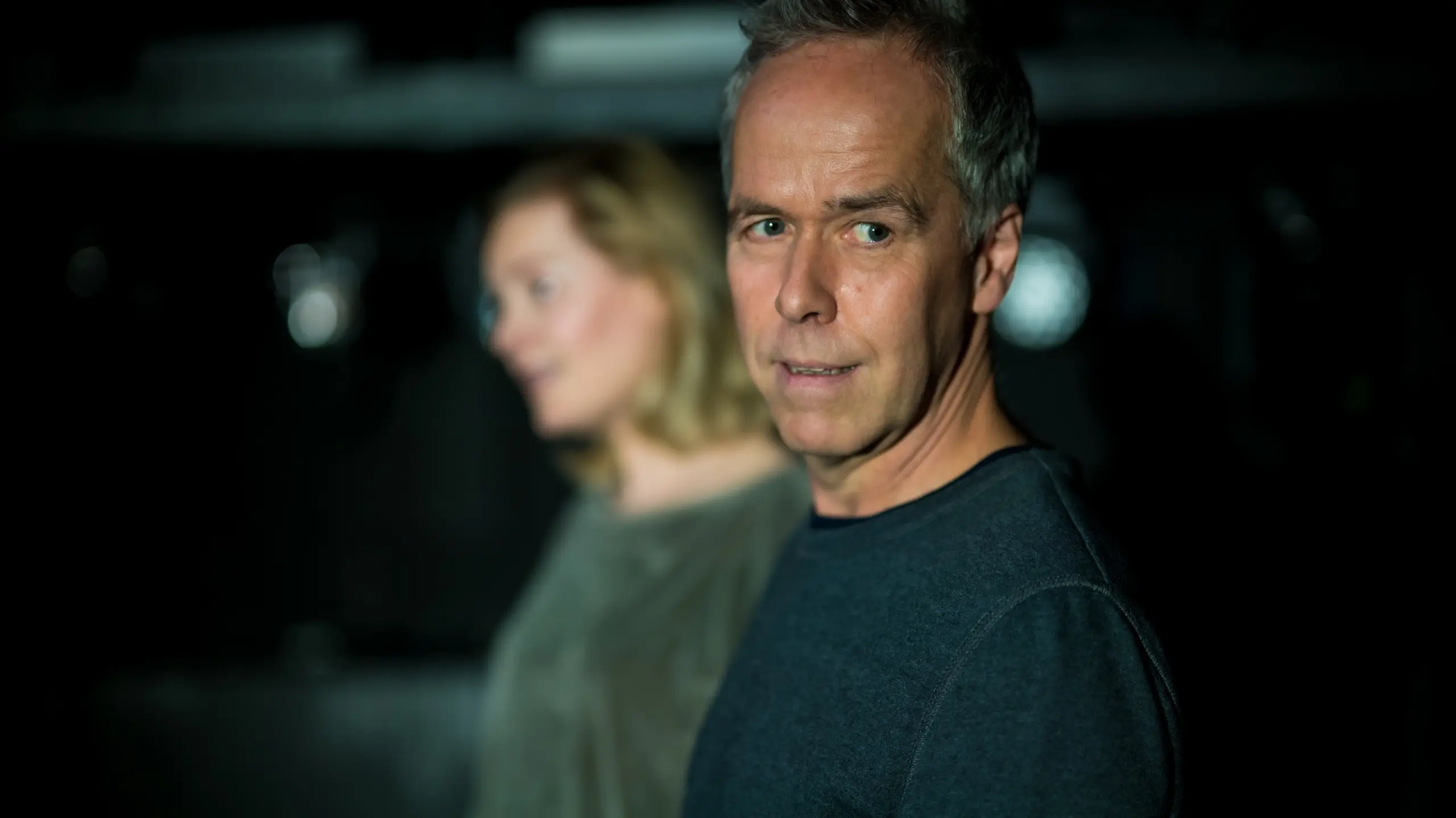 En mann ser rett mot kamera med nøytral mine, mens en kvinne står bak ham i uskarpt fokus, begge i mørke klær.