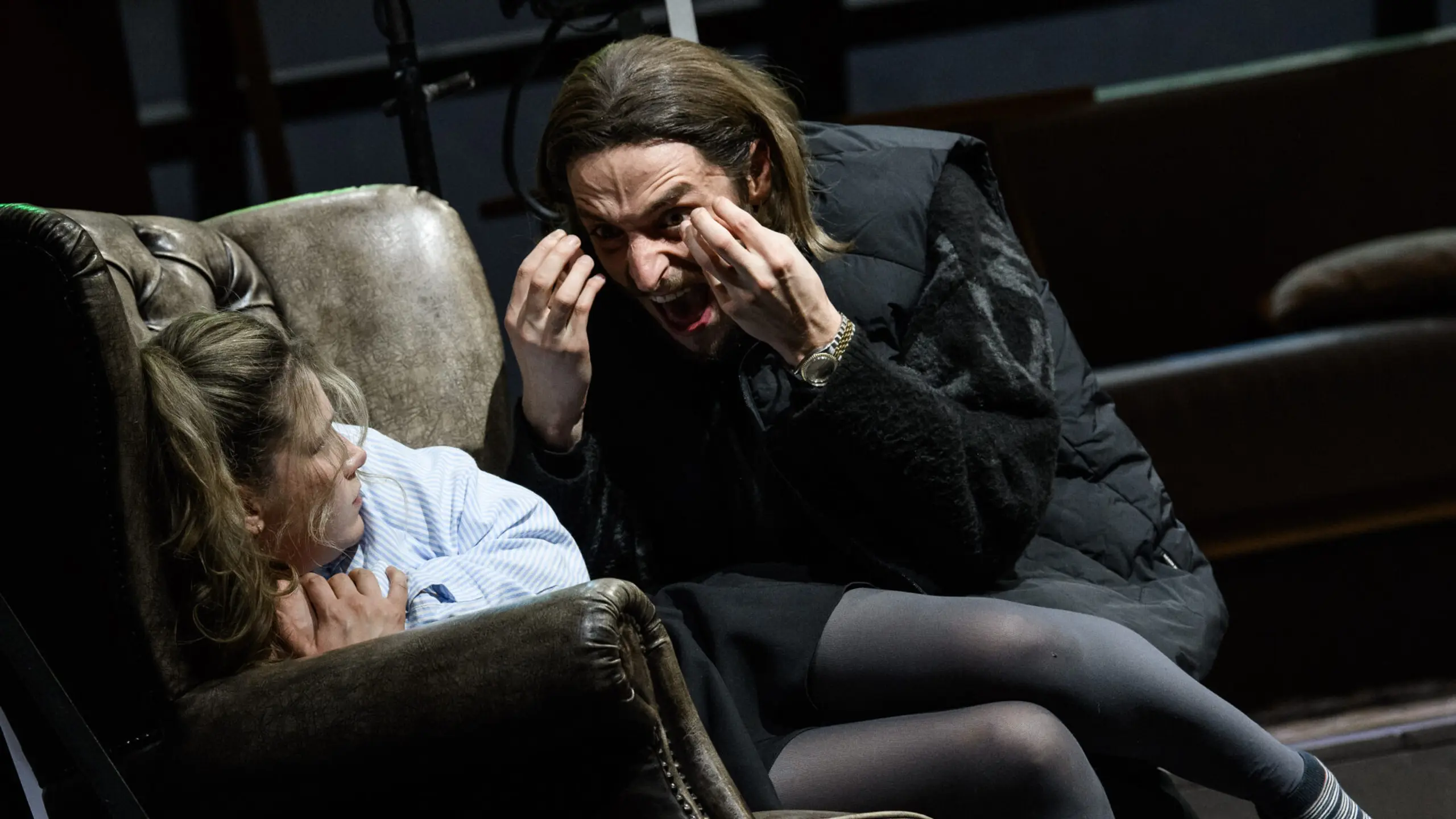 Mann med skulderlangt hår gestikulerer intenst mot en ung kvinne som ligger sammenkrøpet i en skinnlenestol.