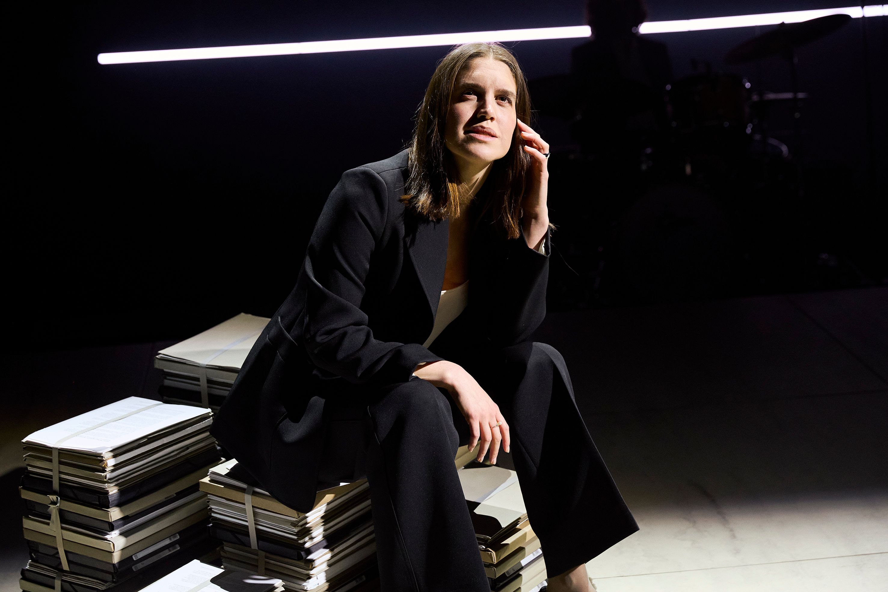 En kvinne i svart dress sitter på en bunke med dokumenter, med hånden til kinnet.