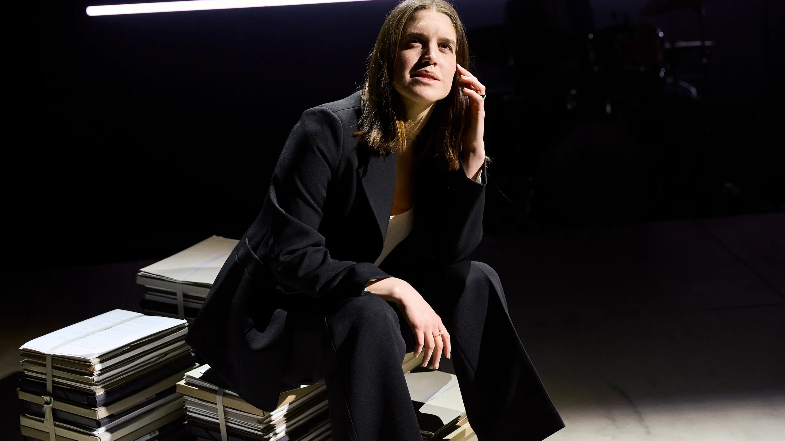 En kvinne i svart dress sitter på en bunke med dokumenter, med hånden til kinnet.