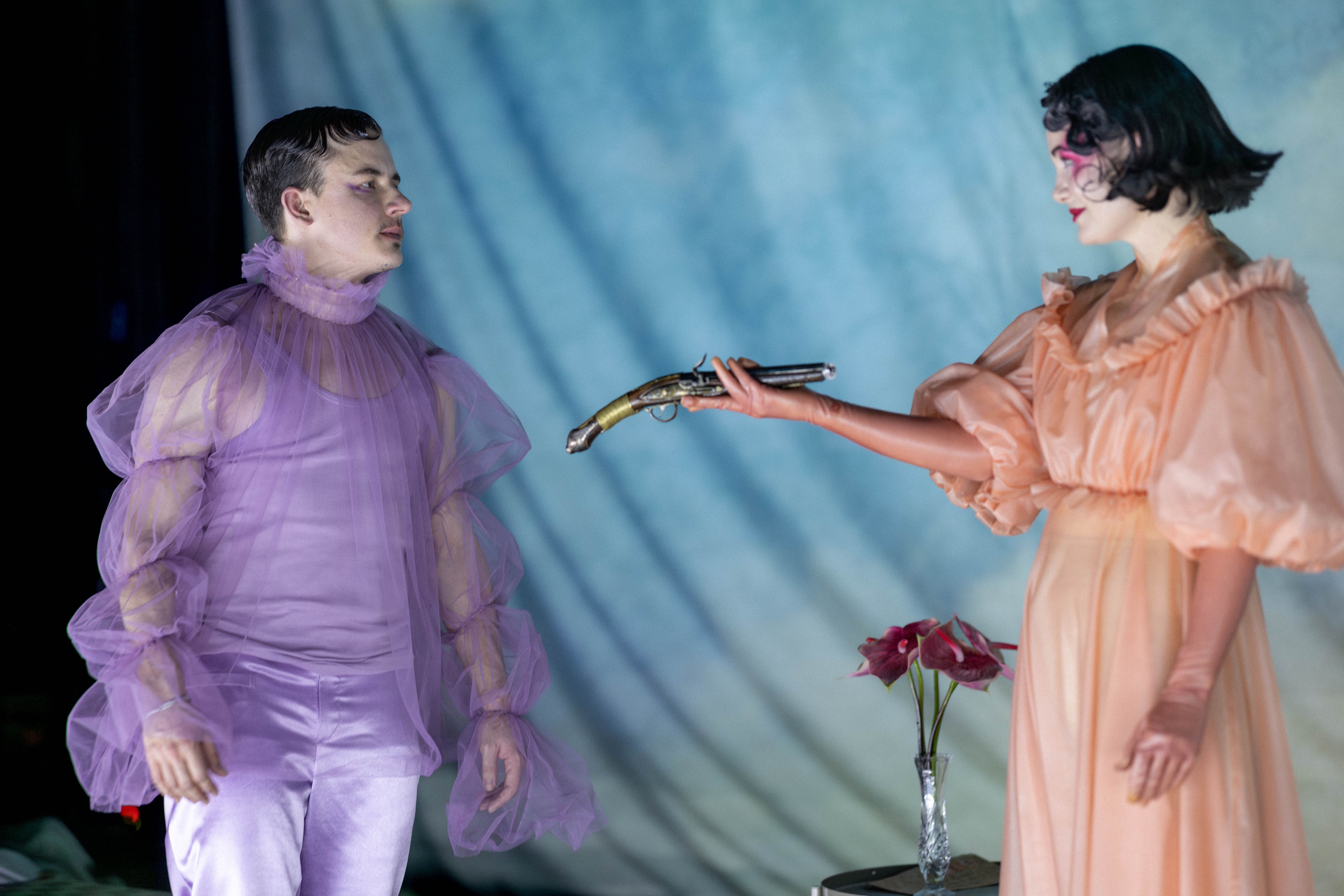 Person i ferskenfarget kjole holder en gammel pistol rettet mot en person i lilla tyllkostyme
