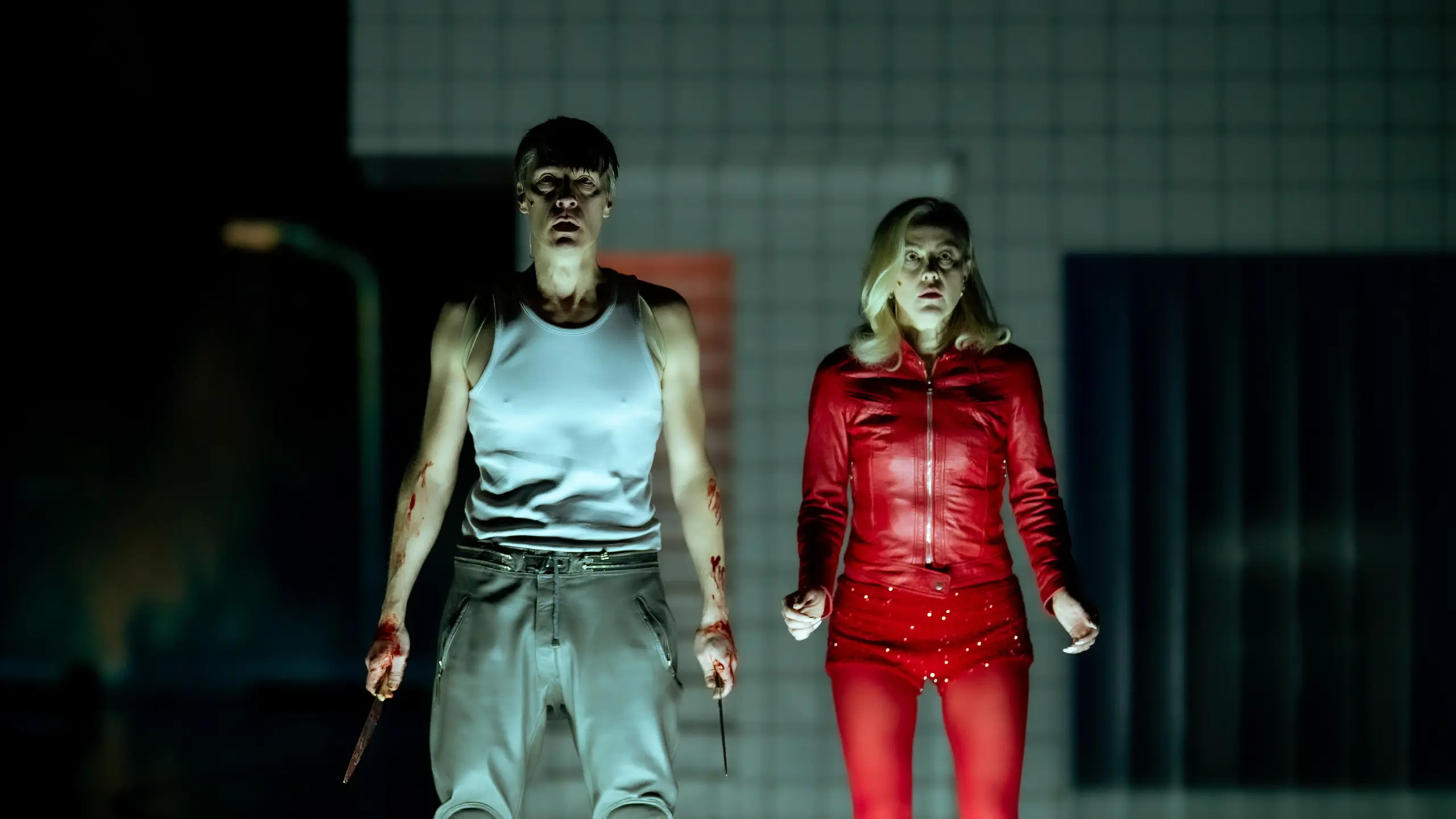 To blodige skuespillere stirrer fremover, den ene i rød drakt og den andre i hvit singlet med dolker i hendene