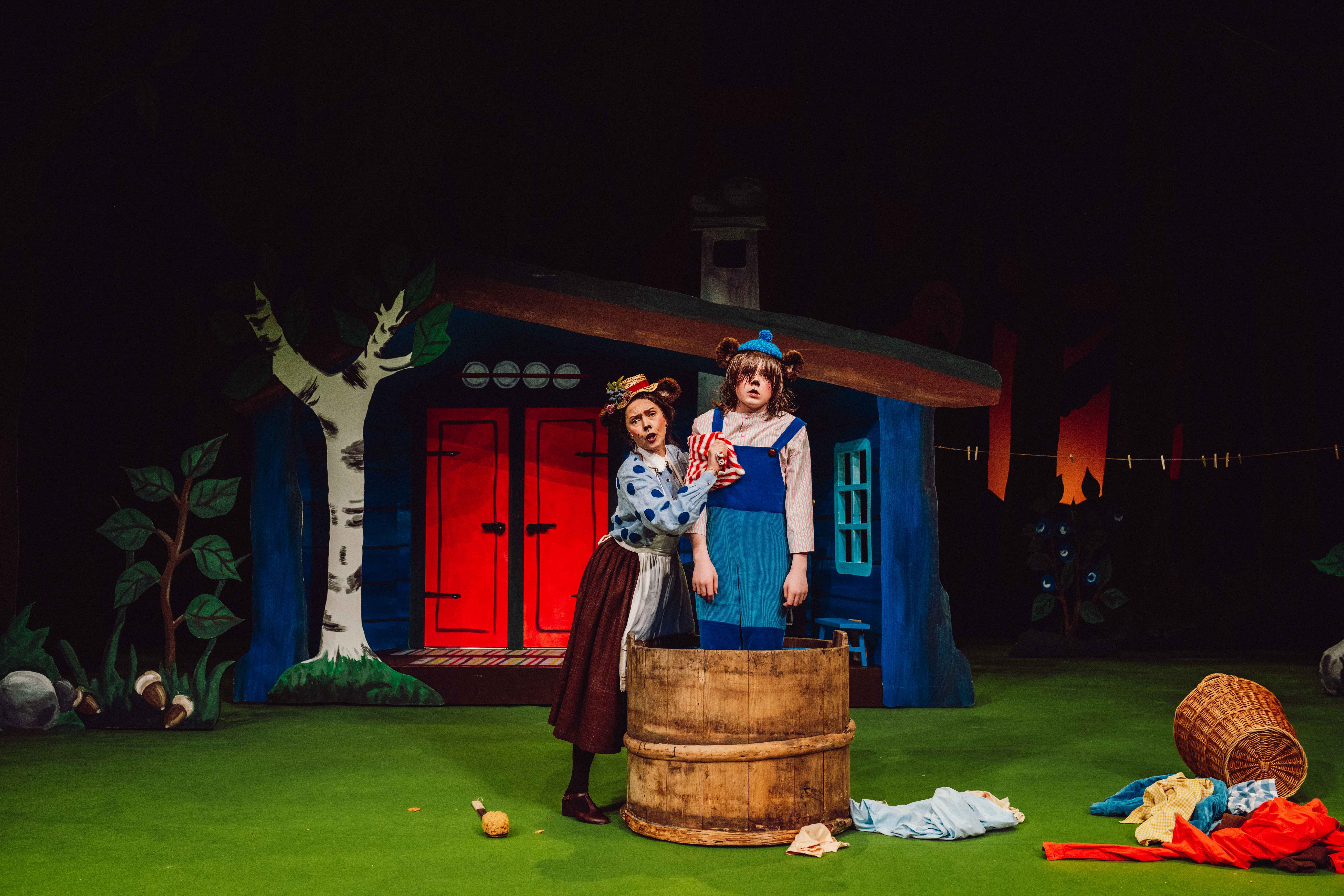 En bjørnemor vasker en bjørneunge i en stor trekum foran et hus med røde dører.