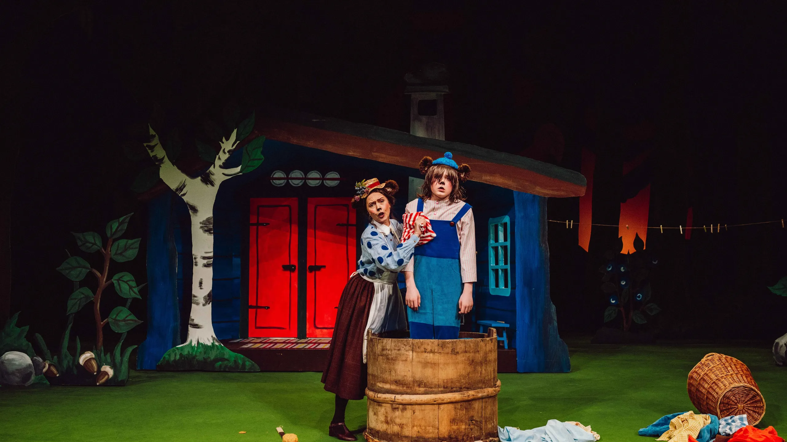 En bjørnemor vasker en bjørneunge i en stor trekum foran et hus med røde dører.