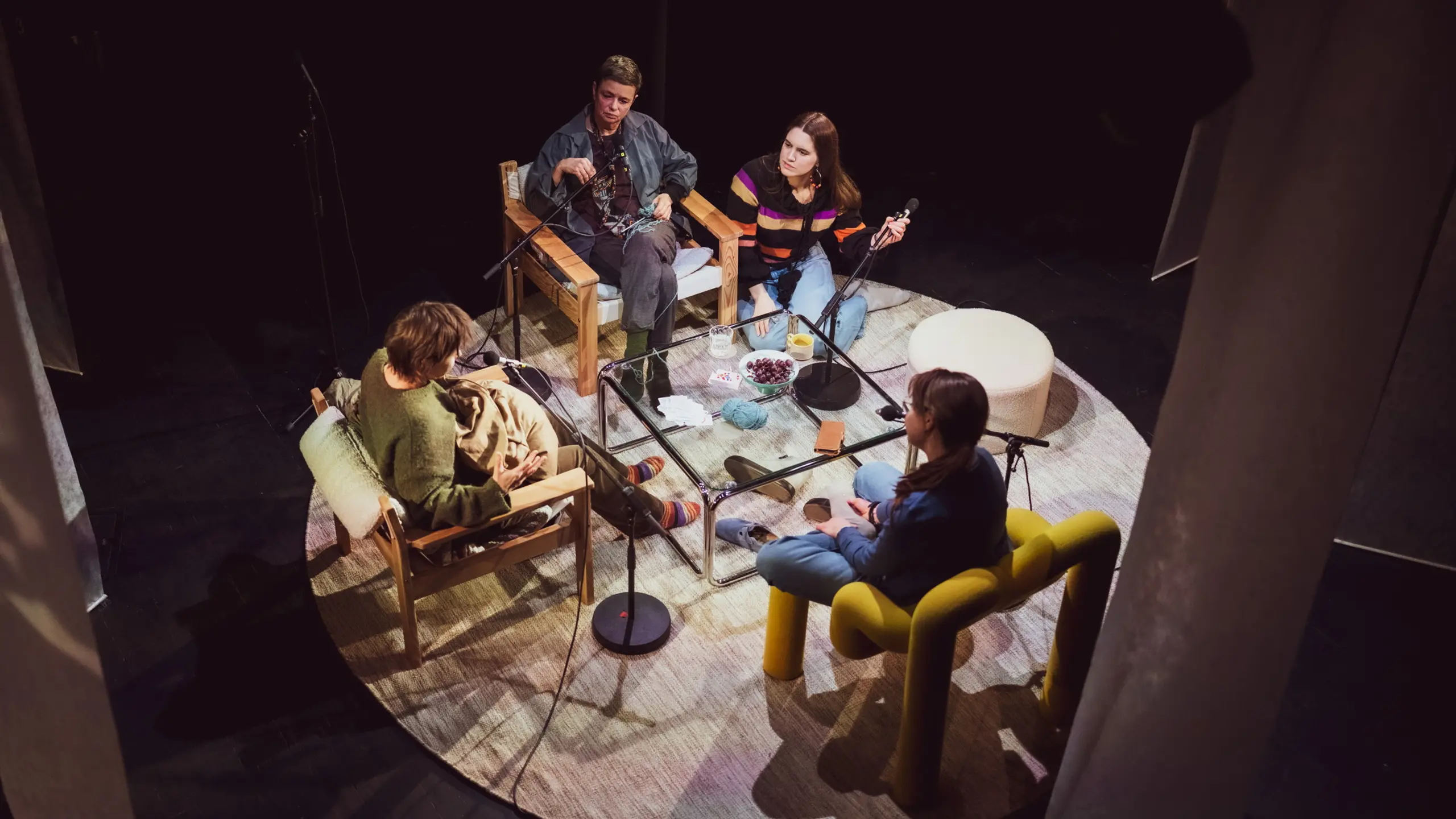 Fire personer i samtalesetting rundt lavt bord med mikrofoner, garn, kopper og fruktfat.