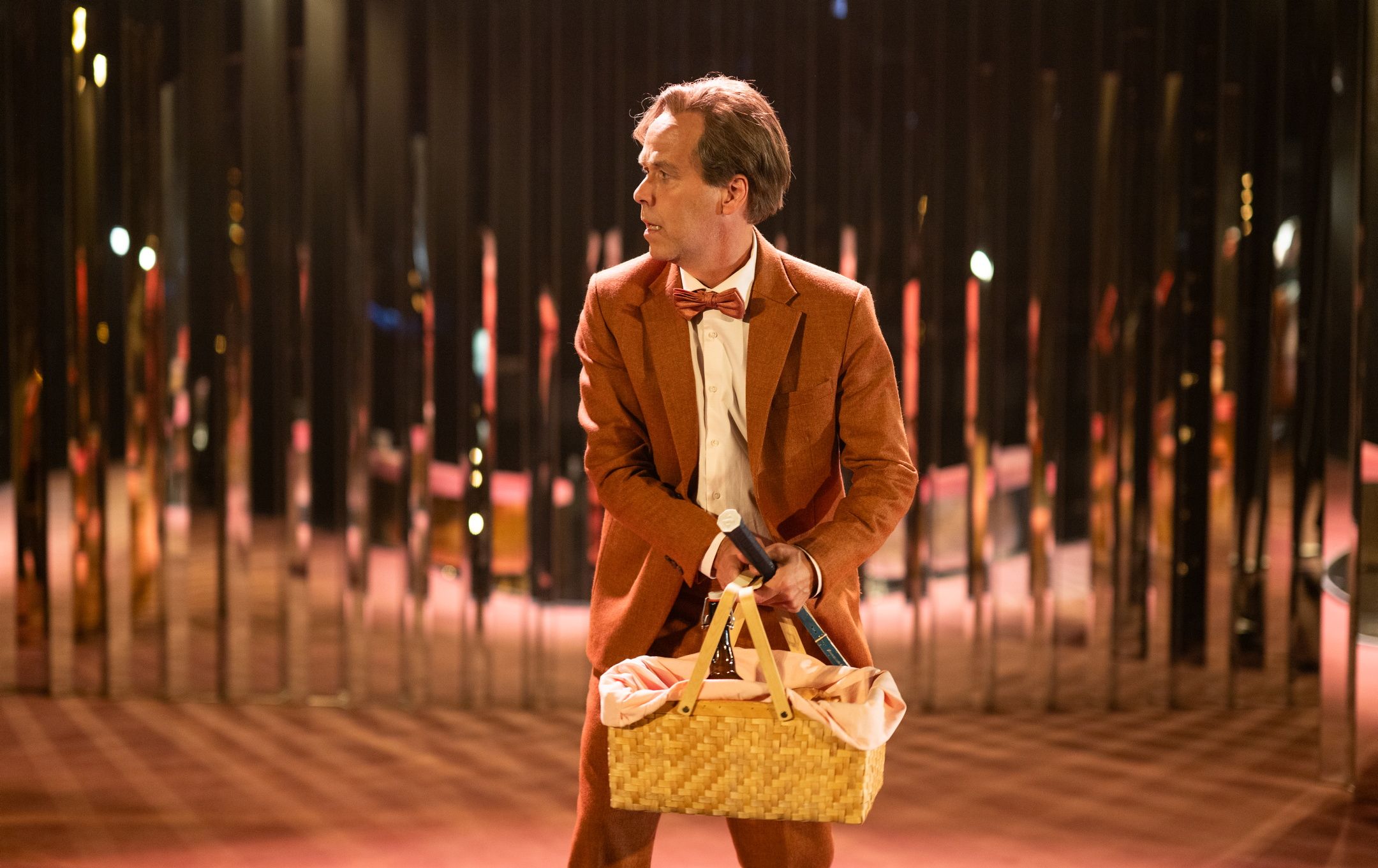 Mann i rød dress og sløyfe holder piknikkurv og mikrofon, ser til siden i rom med speil og varme farger.