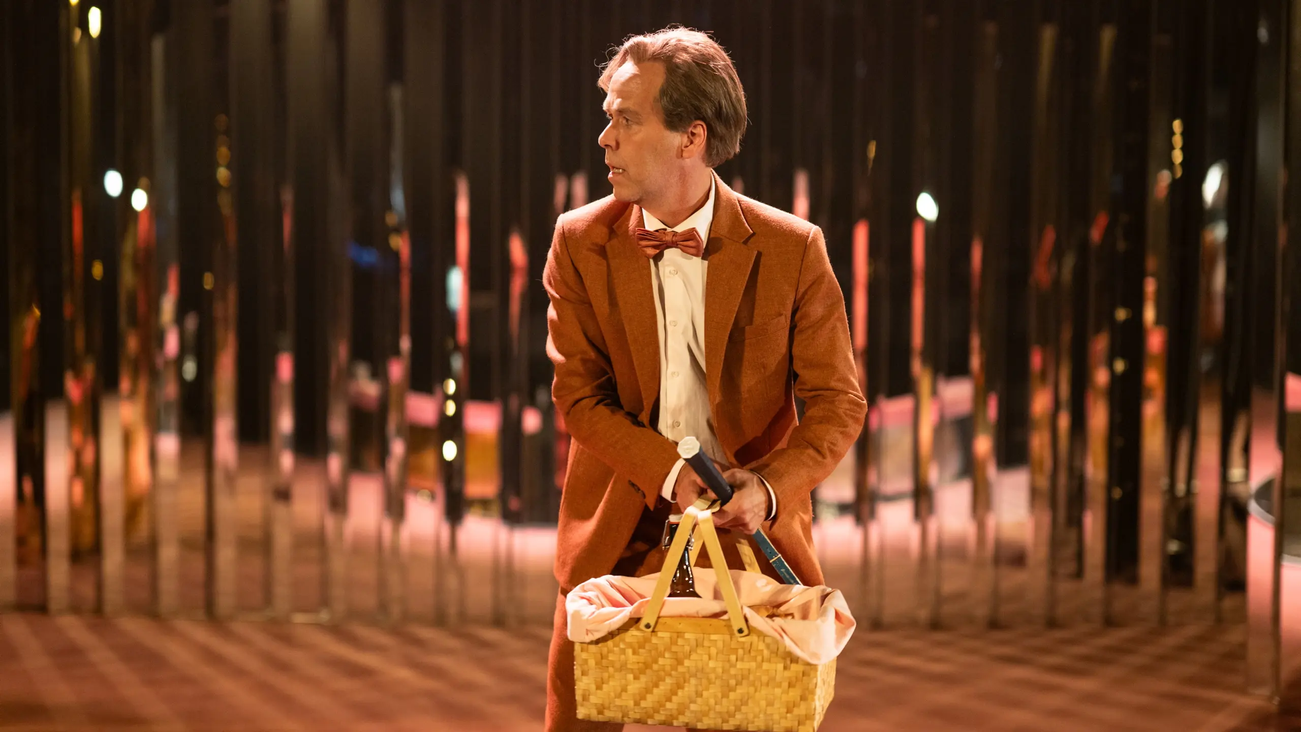 Mann i rød dress og sløyfe holder piknikkurv og mikrofon, ser til siden i rom med speil og varme farger.