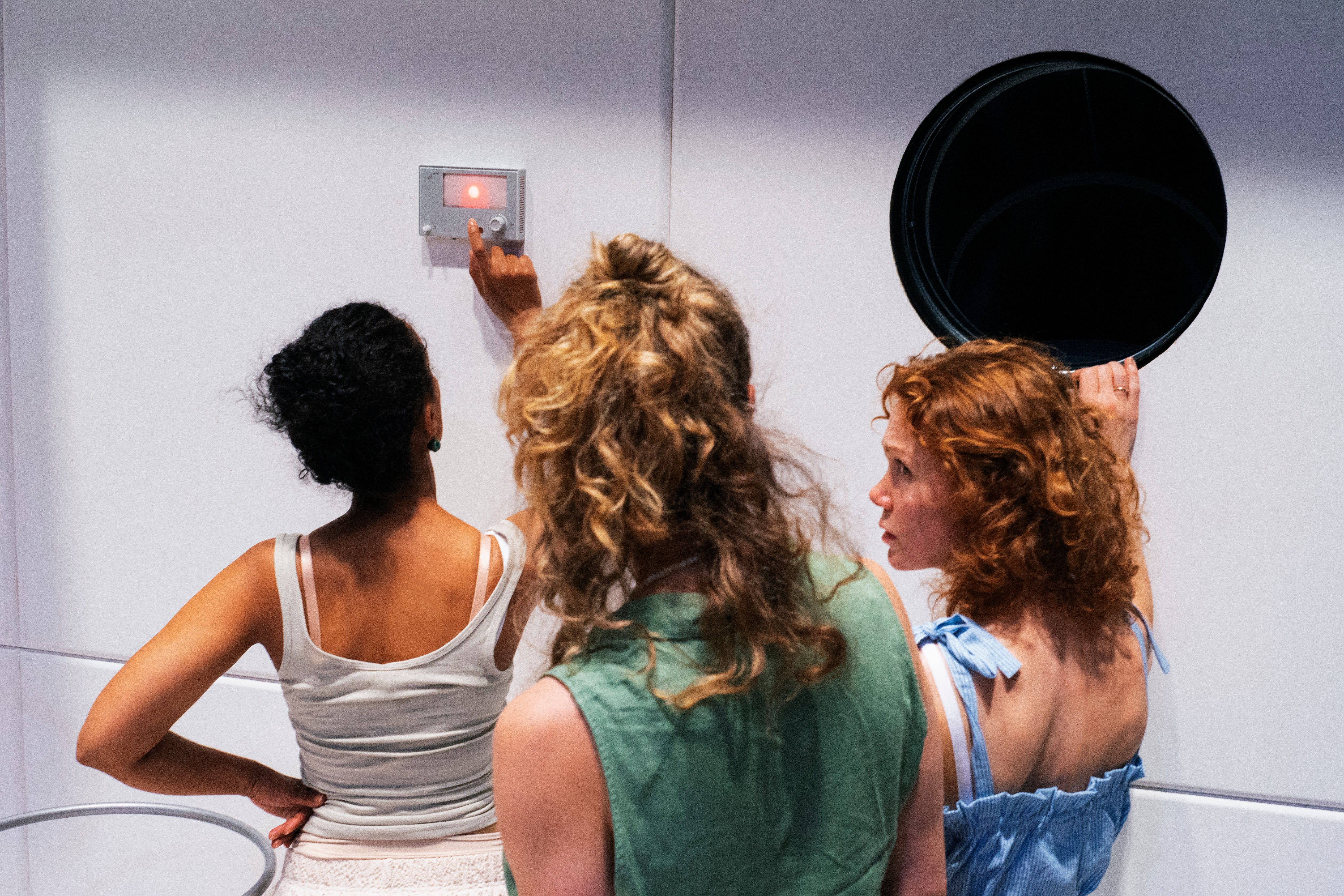 Tre kvinner, sett bakfra, ser på en veggmontert enhet med rødt lys, én peker på knappen.