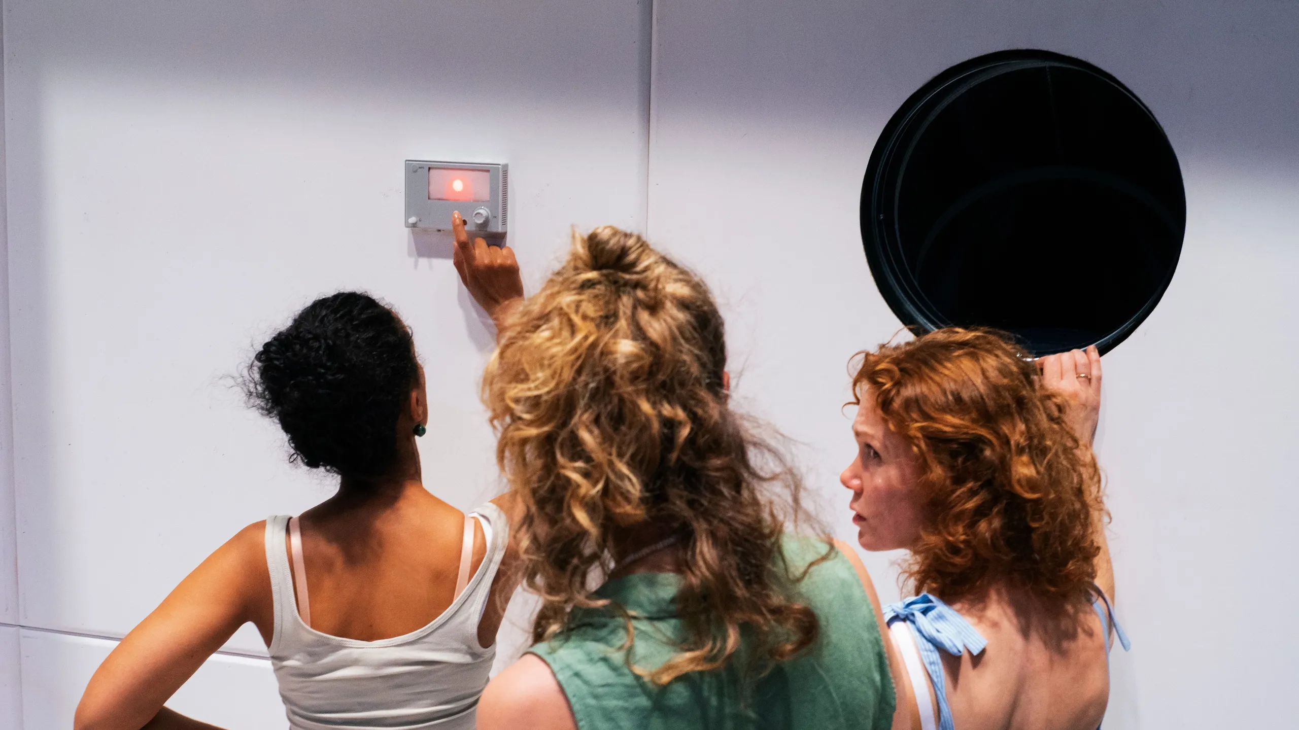 Tre kvinner, sett bakfra, ser på en veggmontert enhet med rødt lys, én peker på knappen.