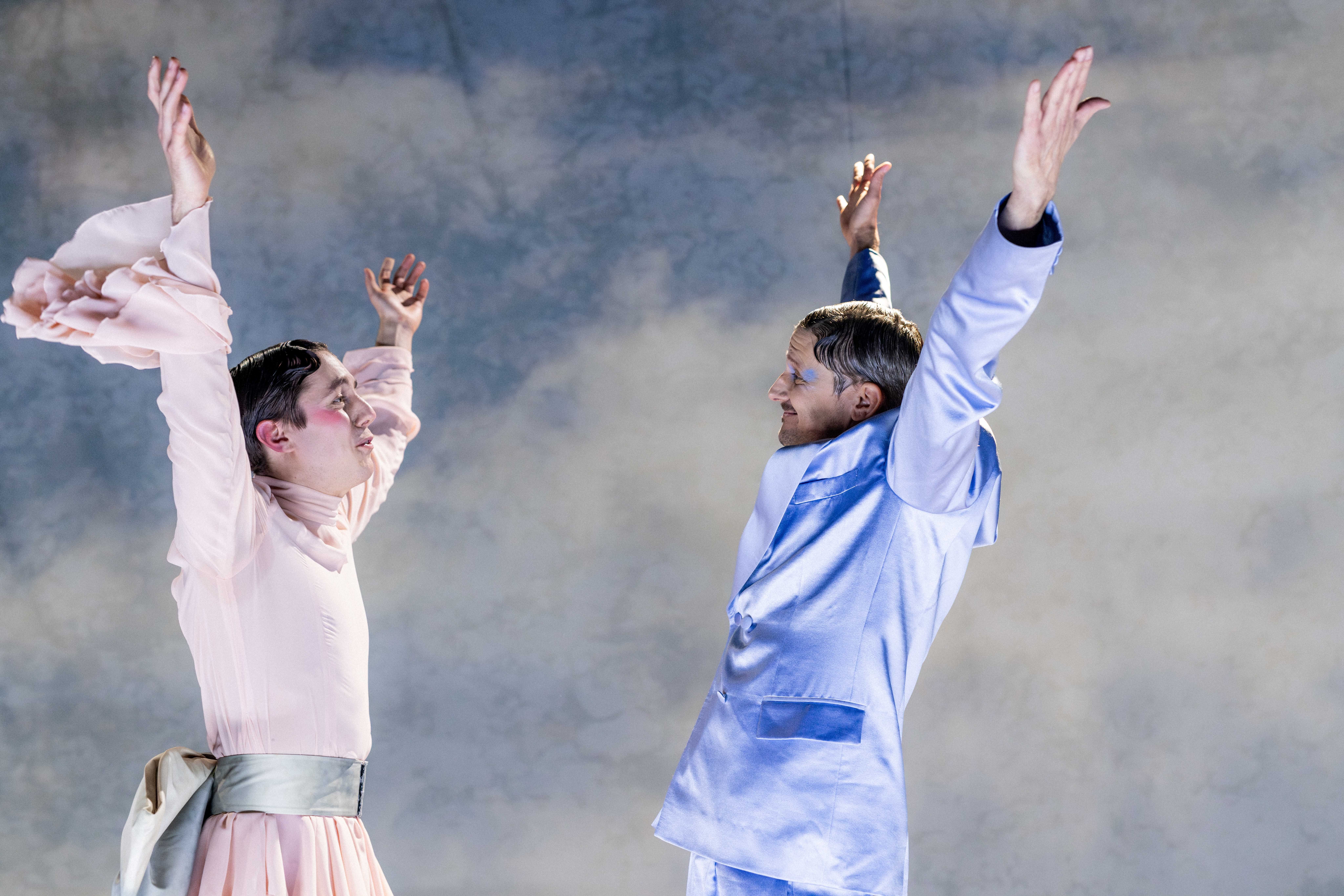 To personer i pastellfargede kostymer står vendt mot hverandre med armene løftet i en teatralsk gest