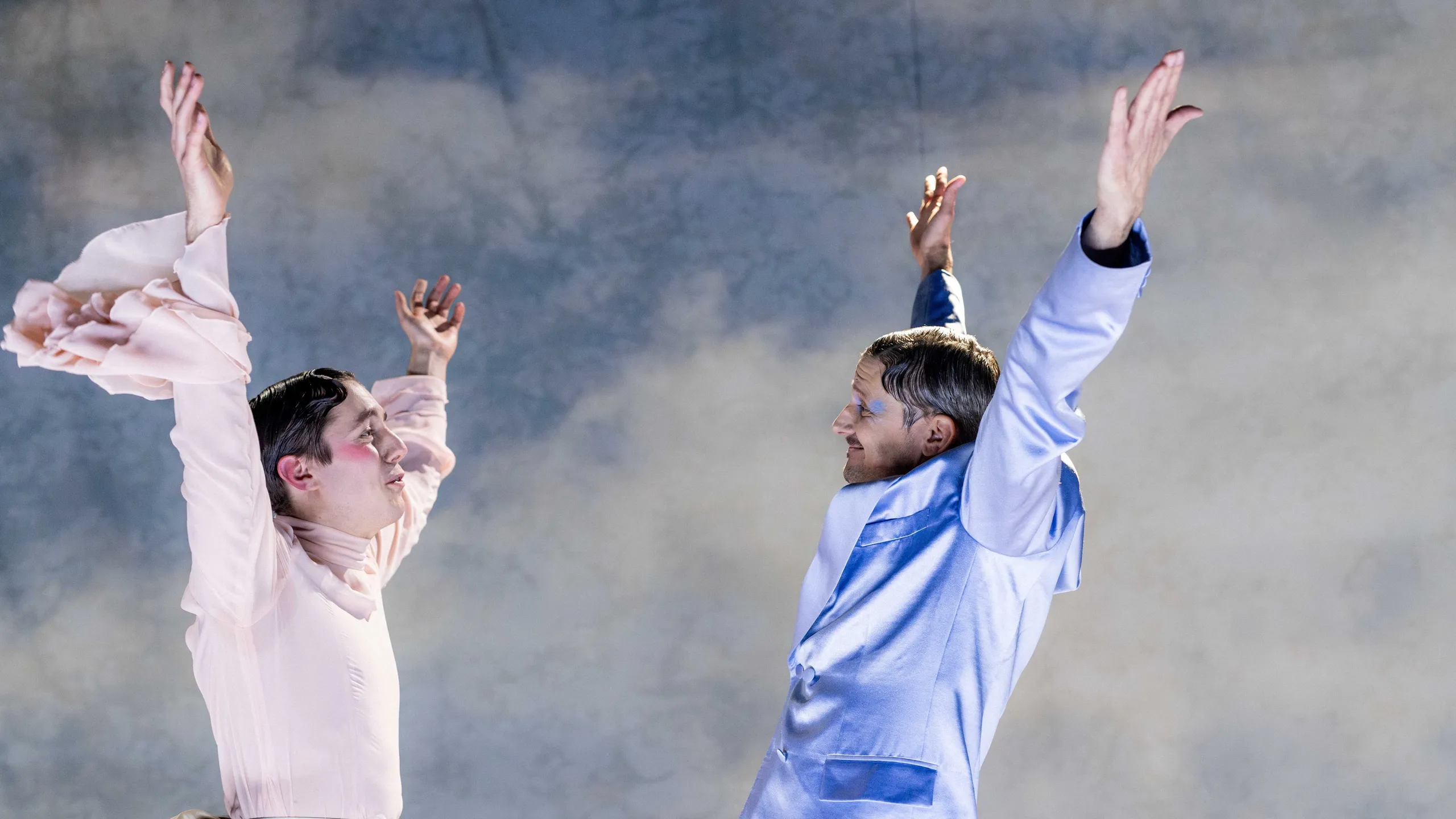 To personer i pastellfargede kostymer står vendt mot hverandre med armene løftet i en teatralsk gest