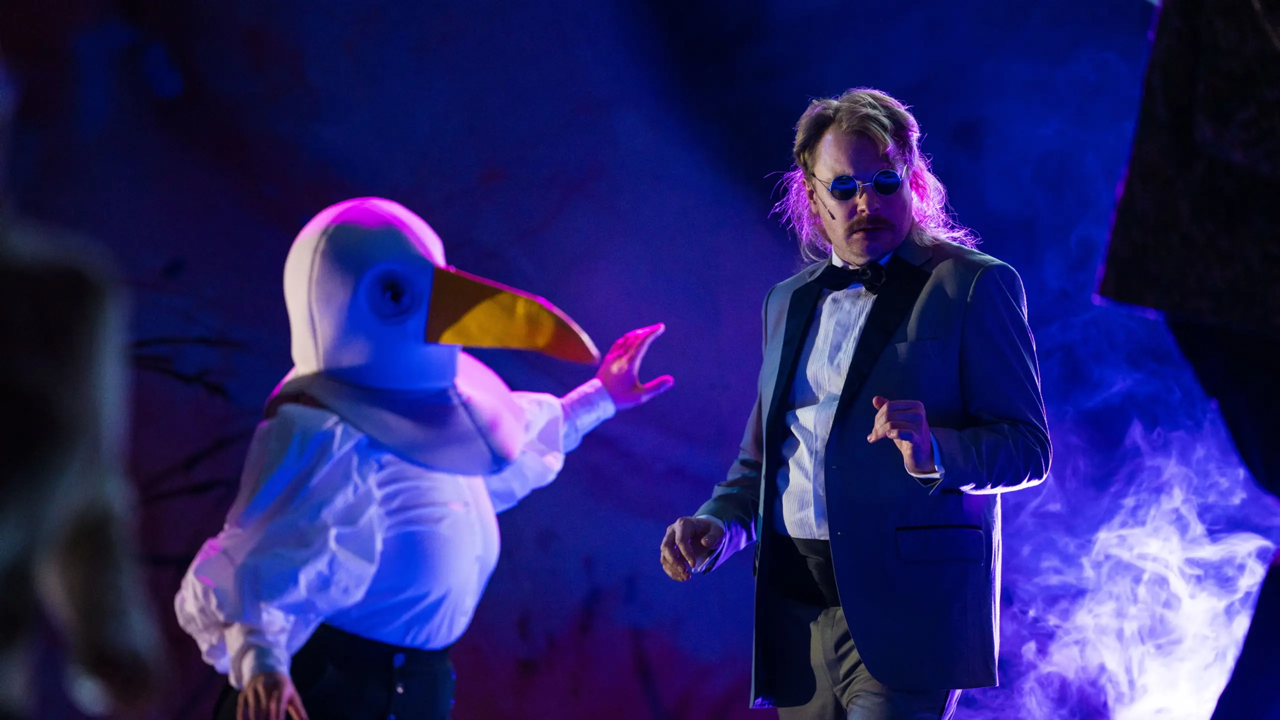 Person i måkekostyme strekker arm mot en person i smoking og blå briller under lilla scenelys.