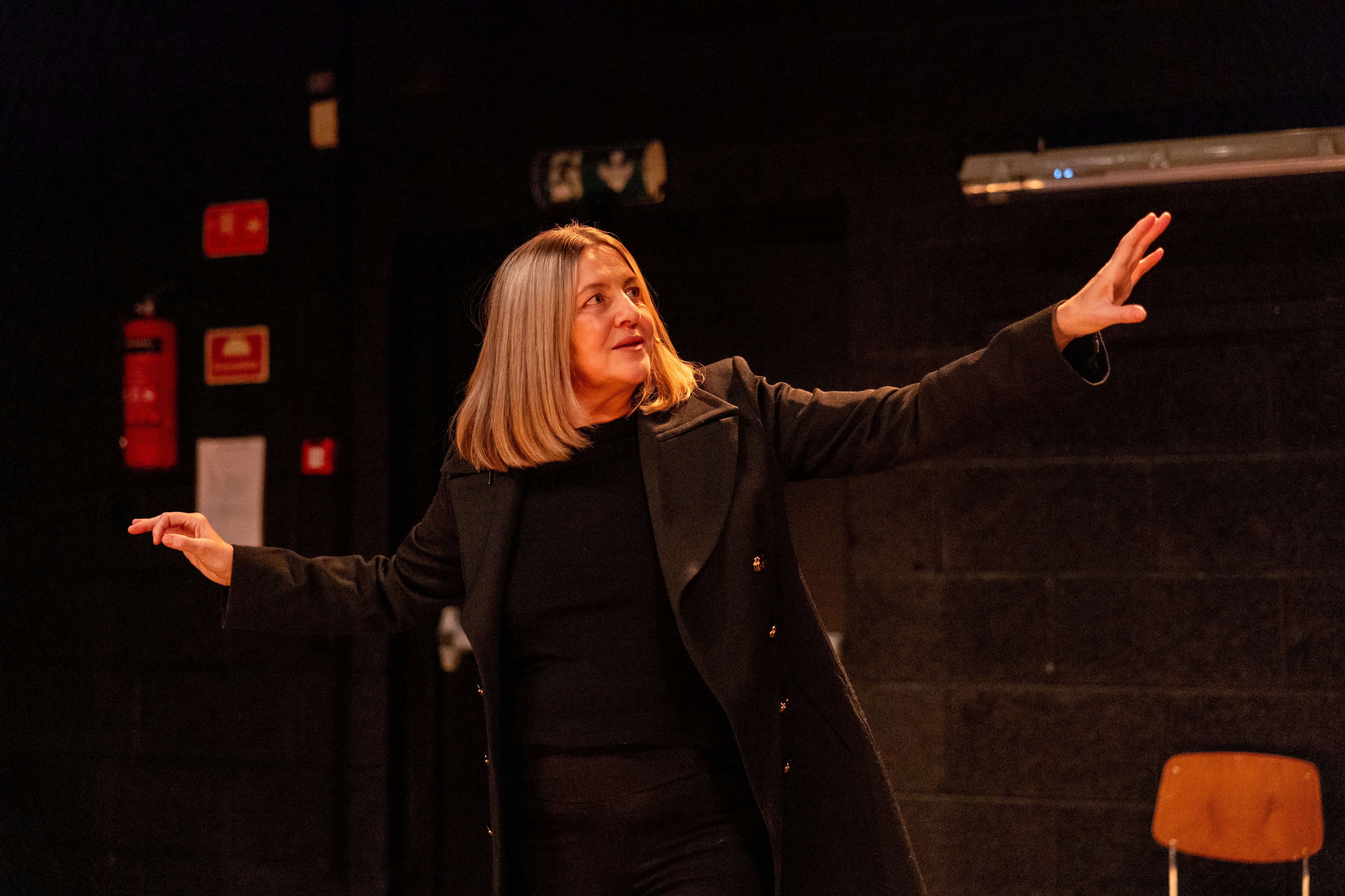 Kvinne med blondt hår i sort antrekk og frakk står med armene løftet foran en mørk vegg og en tom stol.