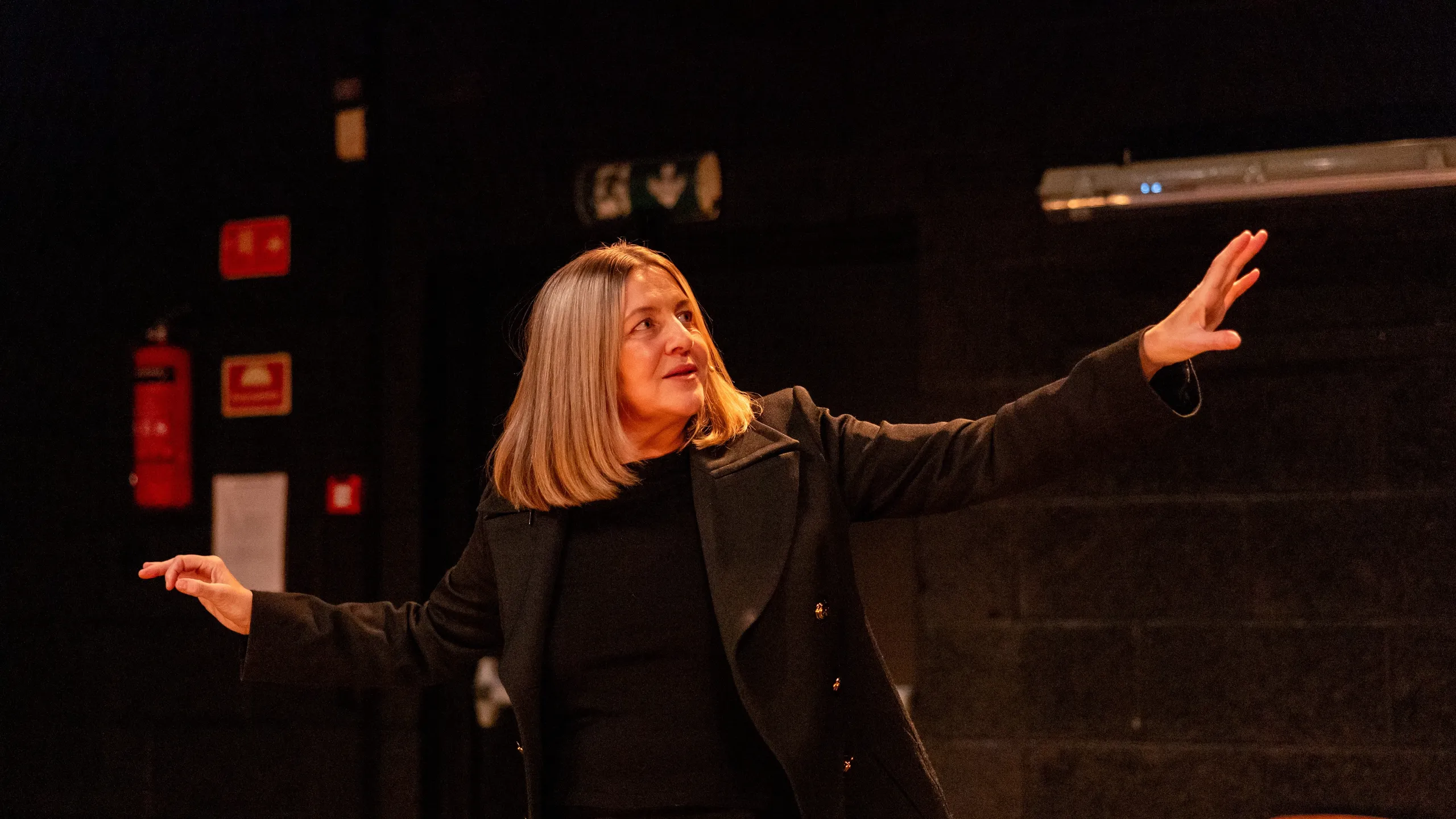 Kvinne med blondt hår i sort antrekk og frakk står med armene løftet foran en mørk vegg og en tom stol.