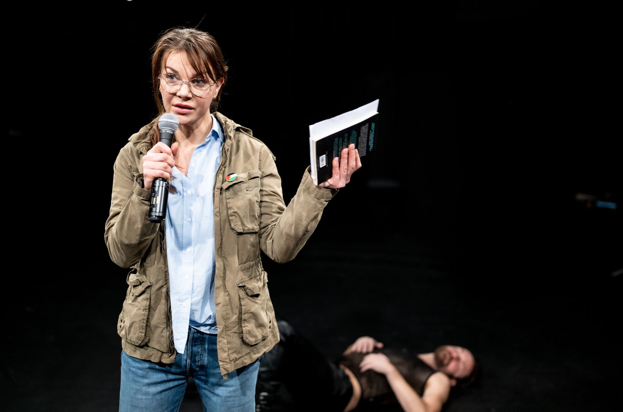 Person med briller holder mikrofon og bok mens en annen person ligger på scenegulvet bak.