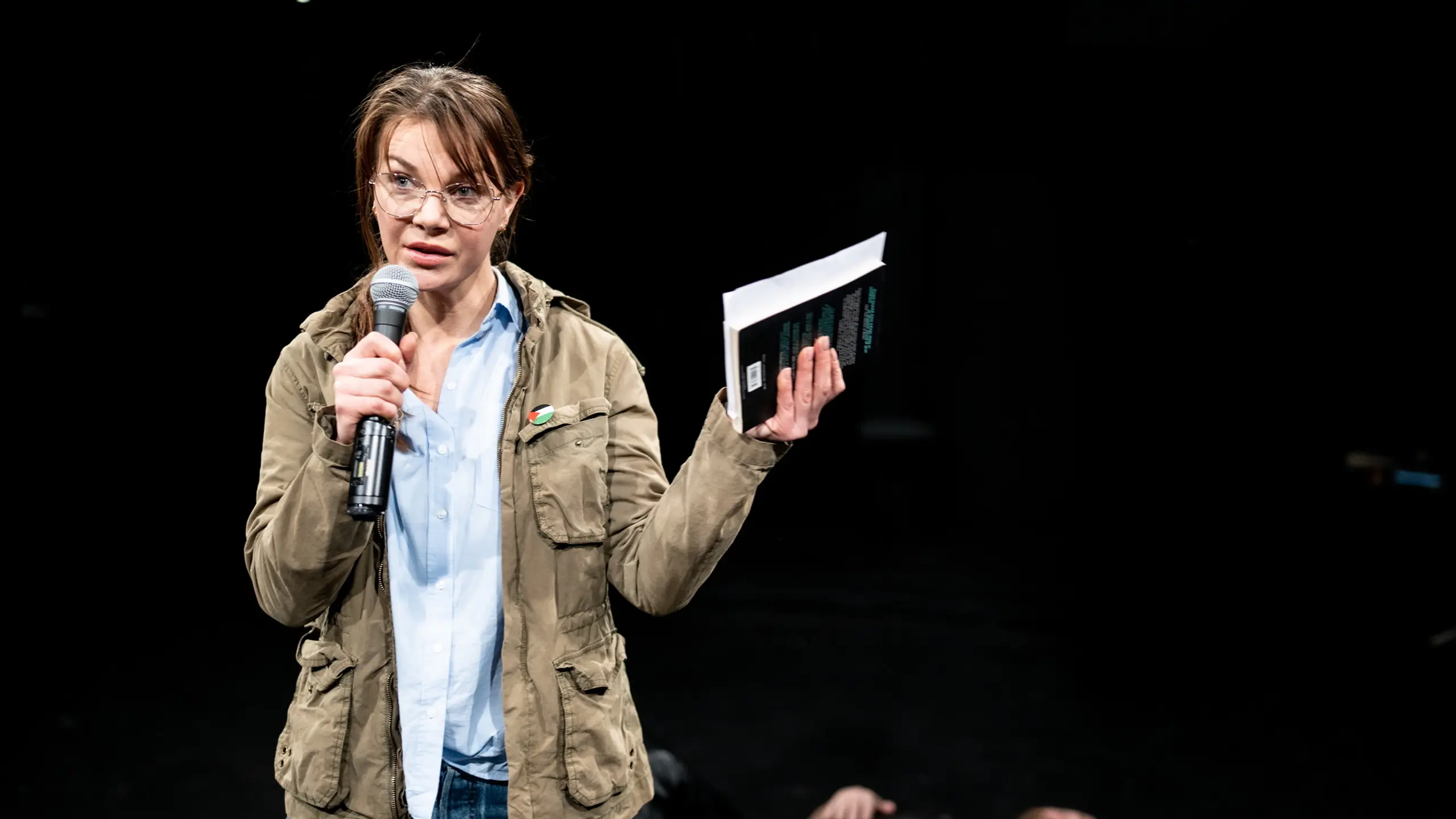 Person med briller holder mikrofon og bok mens en annen person ligger på scenegulvet bak.