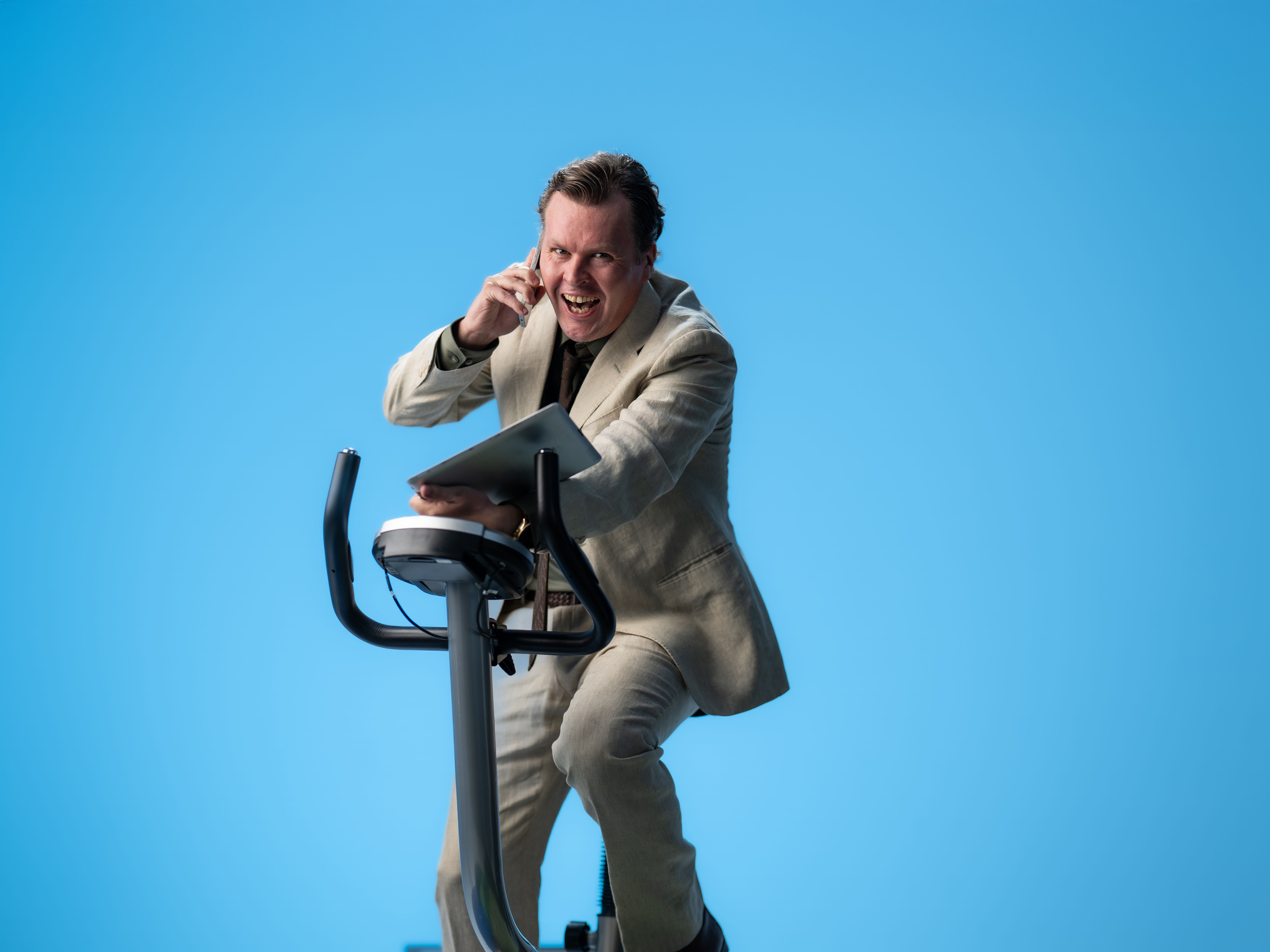 Smilende mann i dress som snakker i telefonen på ergometersykkel.