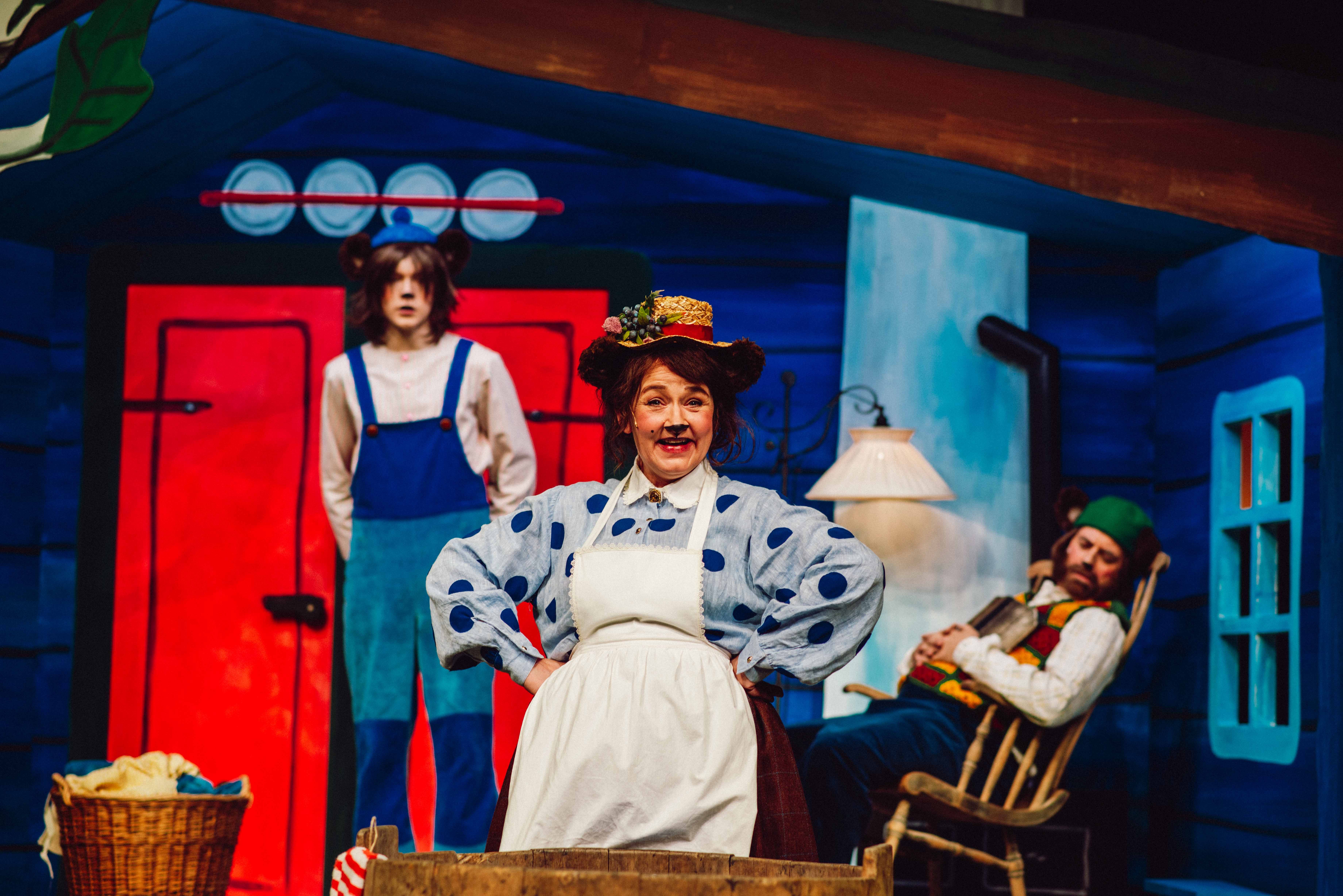 En kvinne utkledd som bjørn står smilende foran et vaskebrett, mens en person i blå snekkerbukse og en annen i gyngestol er i bakgrunnen.