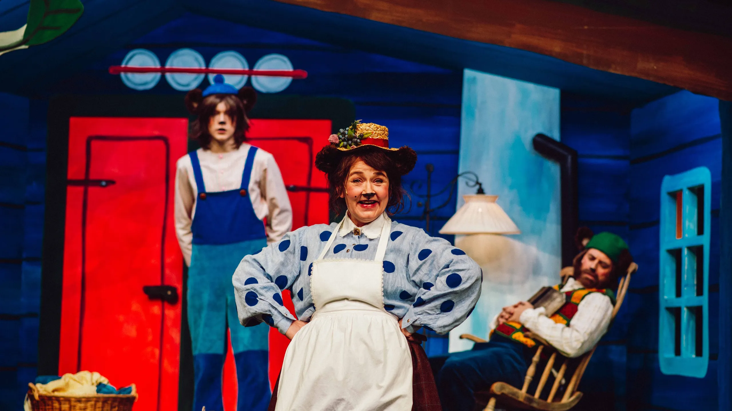 En kvinne utkledd som bjørn står smilende foran et vaskebrett, mens en person i blå snekkerbukse og en annen i gyngestol er i bakgrunnen.